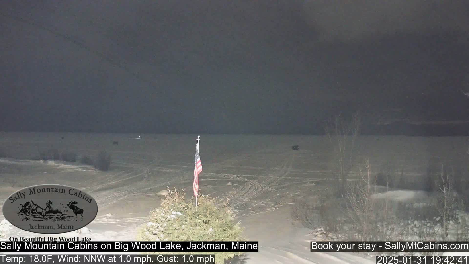A nighttime view of a snow-covered lake with a small American flag in the foreground, under a dark, overcast sky.