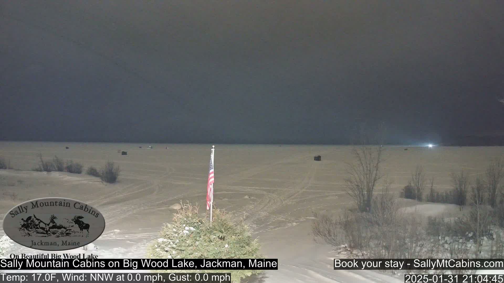 A frozen lake at night under a dark sky, with a few small structures visible on the ice and a US flag planted in the snow in the foreground.