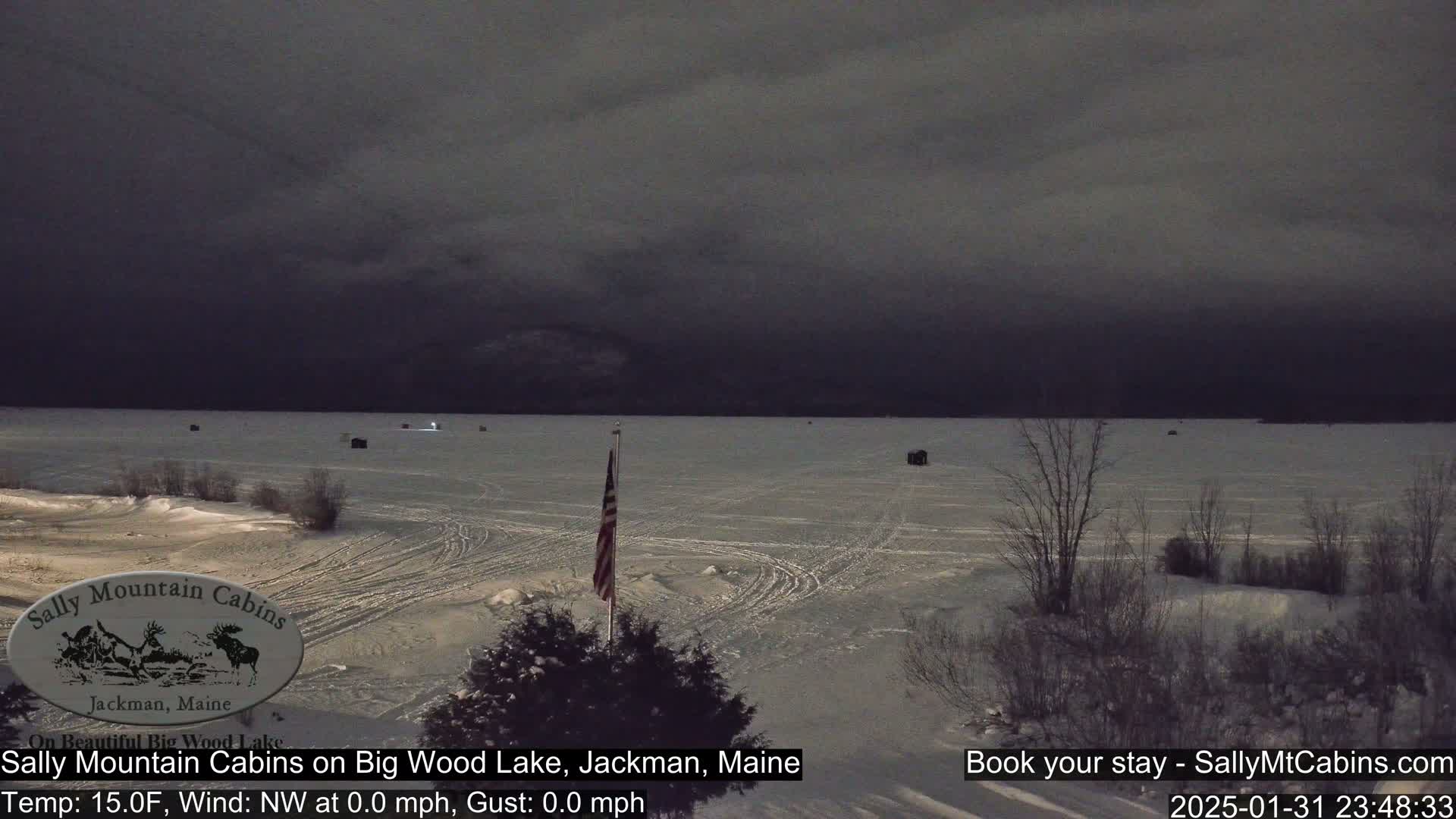 A snow-covered frozen lake is visible under a dark, overcast sky, with several small structures scattered across the ice and a flagpole with an American flag in the foreground.