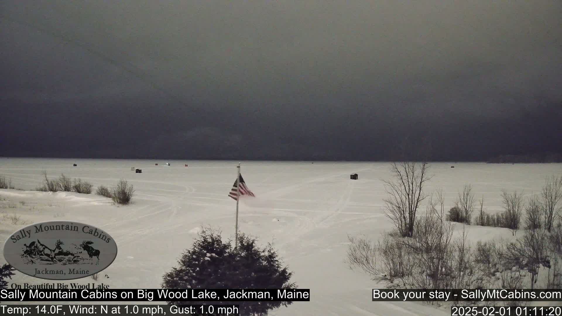 A frozen lake under a dark, overcast sky, with several small structures scattered across the ice and an American flag waving near the foreground.