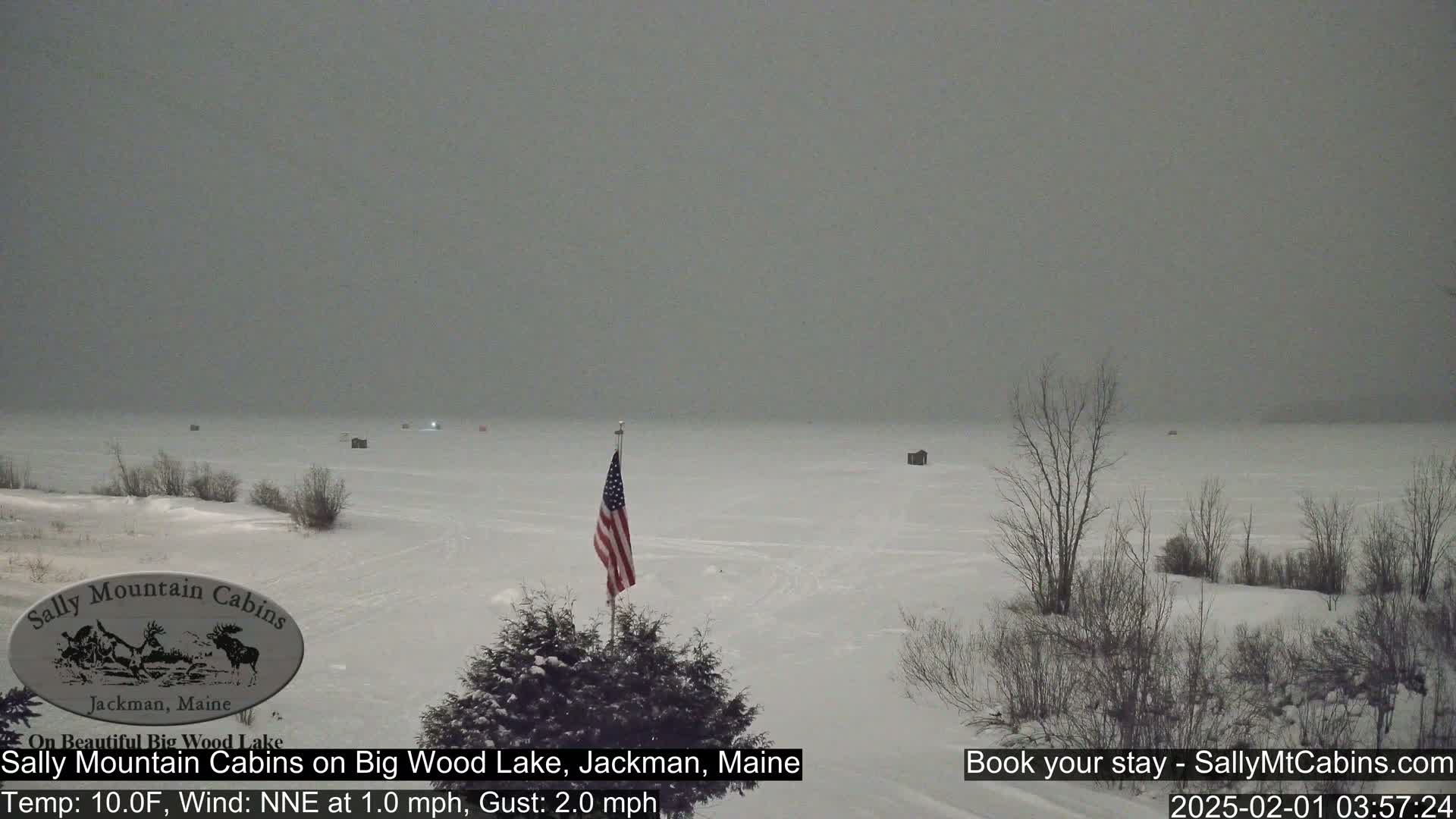 A snow-covered frozen lake is visible under an overcast sky, with a few ice fishing shacks and a small American flag on a pole in the foreground.