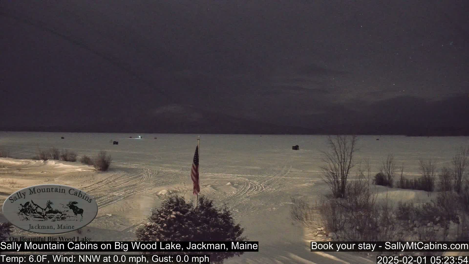 A frozen lake at night under a dark sky, with a few ice fishing shacks visible and an American flag standing near the shoreline.