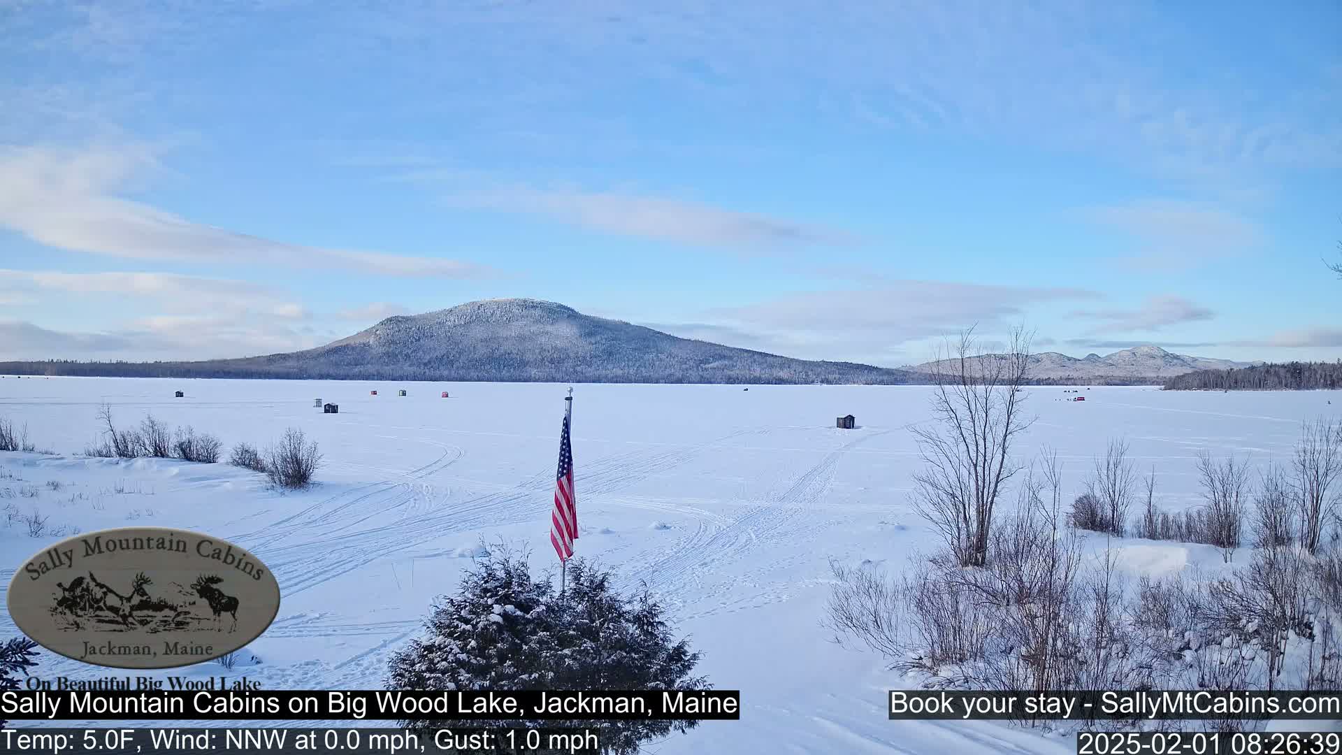 A snow-covered frozen lake stretches out before a treeless hill under a mostly clear blue sky, with several small ice fishing shelters scattered across the ice and an American flag standing near the foreground.