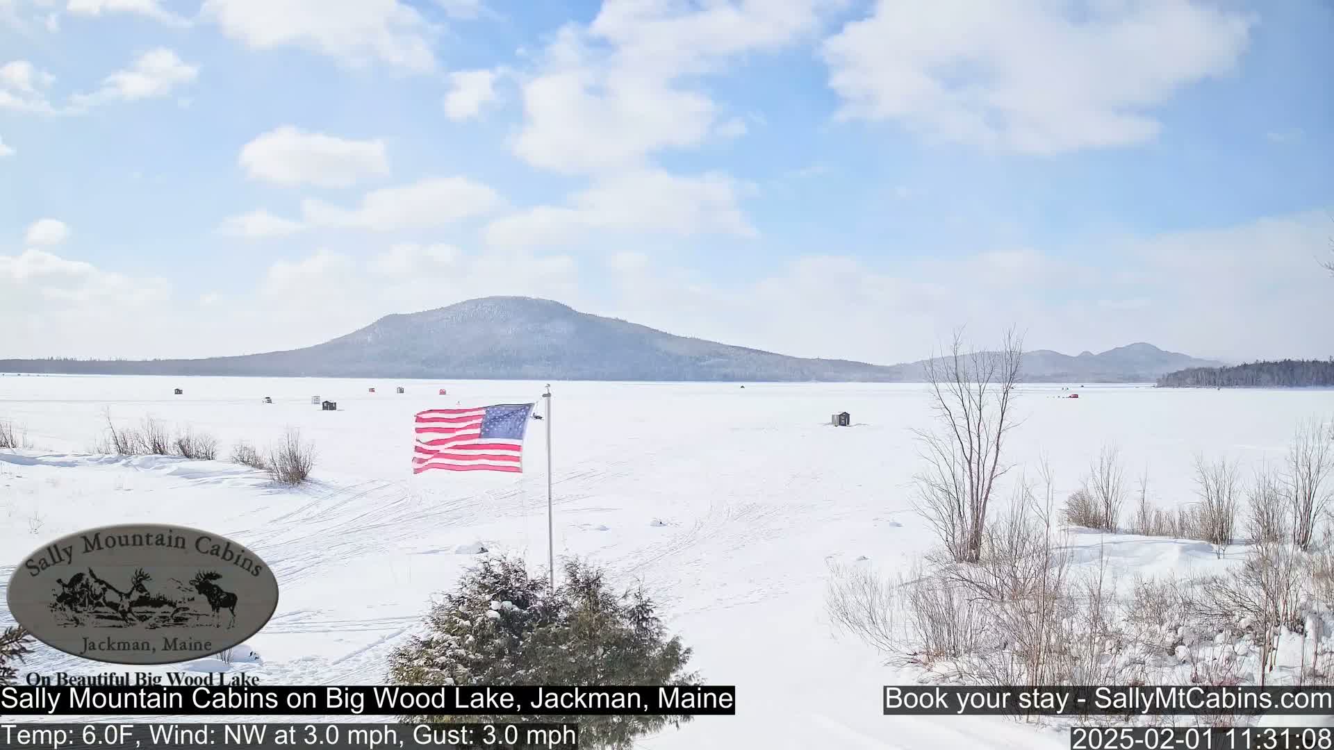 A frozen lake stretches to a snow-covered mountain under a mostly sunny sky, with an American flag waving in the foreground.