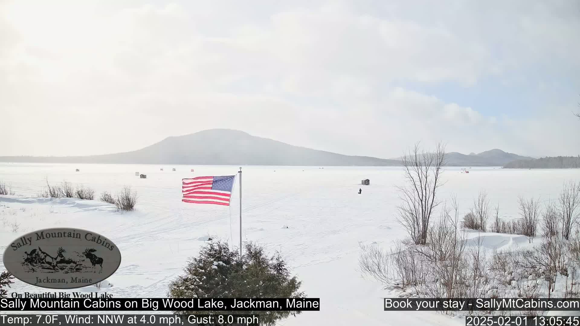A snow-covered frozen lake stretches out to a distant mountain range under a mostly clear sky, with an American flag displayed near the shore.