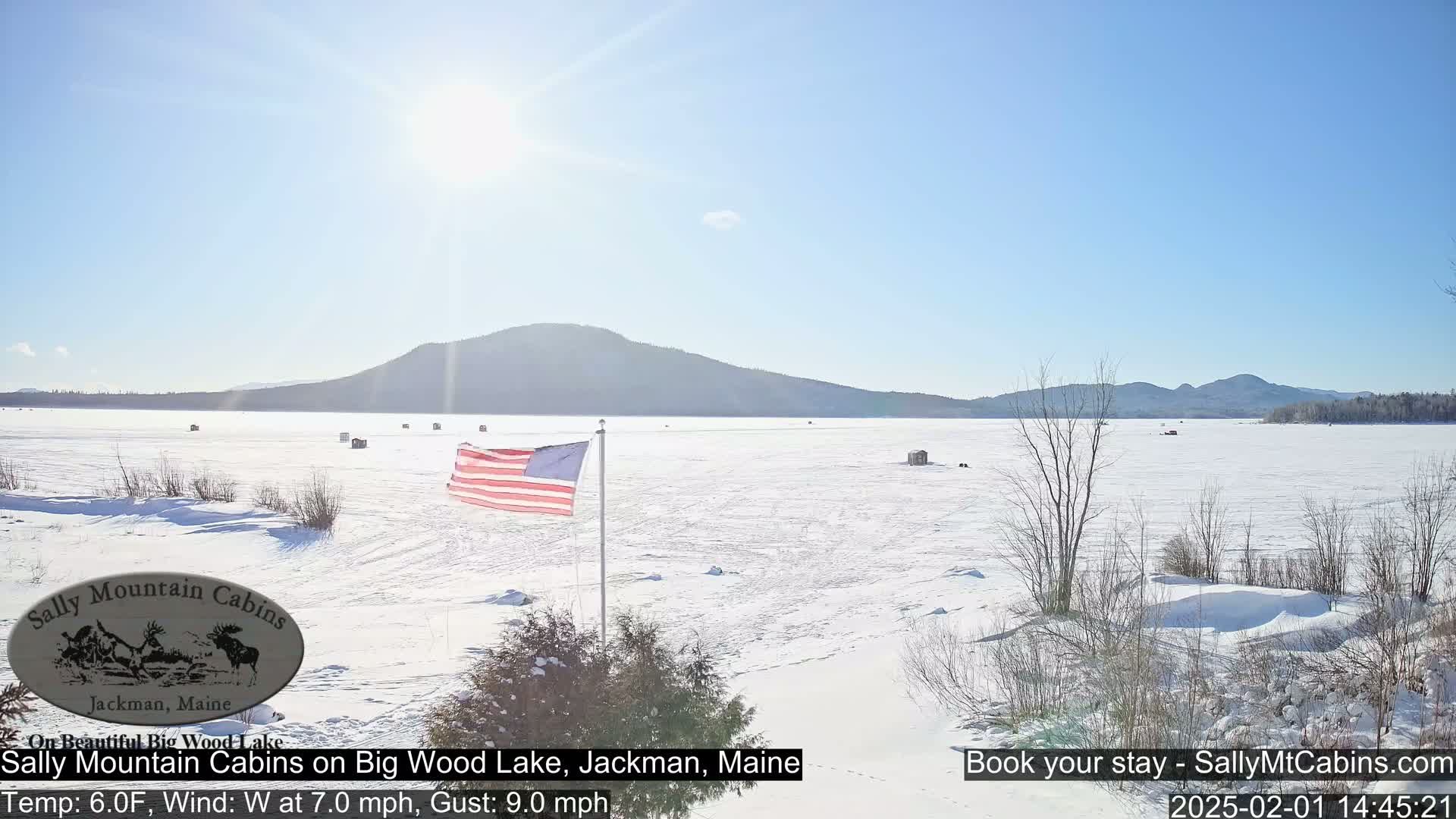 A frozen lake, under a clear sunny sky, is dotted with several ice fishing shelters, and an American flag flies near the shoreline.