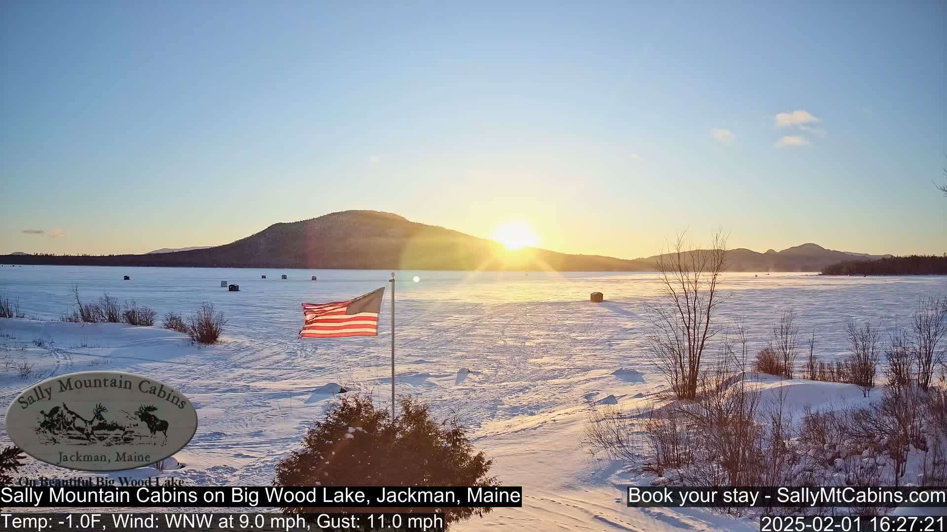 A frozen lake at sunrise, with a snow-covered shoreline, several ice fishing shelters, and an American flag waving in a WNW wind of 9 mph with gusts to 11 mph, and a temperature of -1 degree Fahrenheit.