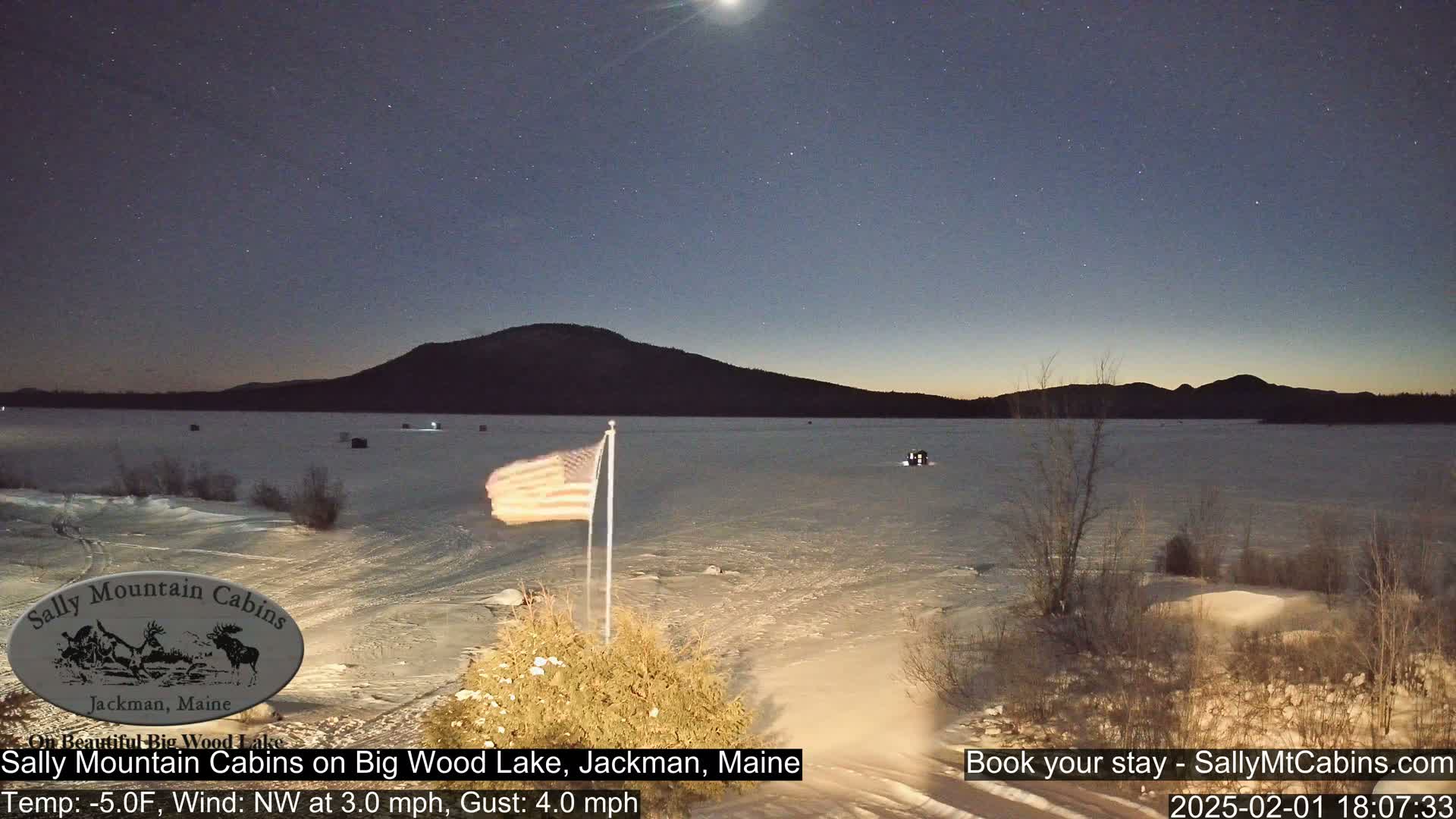 A snow-covered frozen lake is seen at night under a starry sky, with a distant mountain range and an American flag in the foreground.