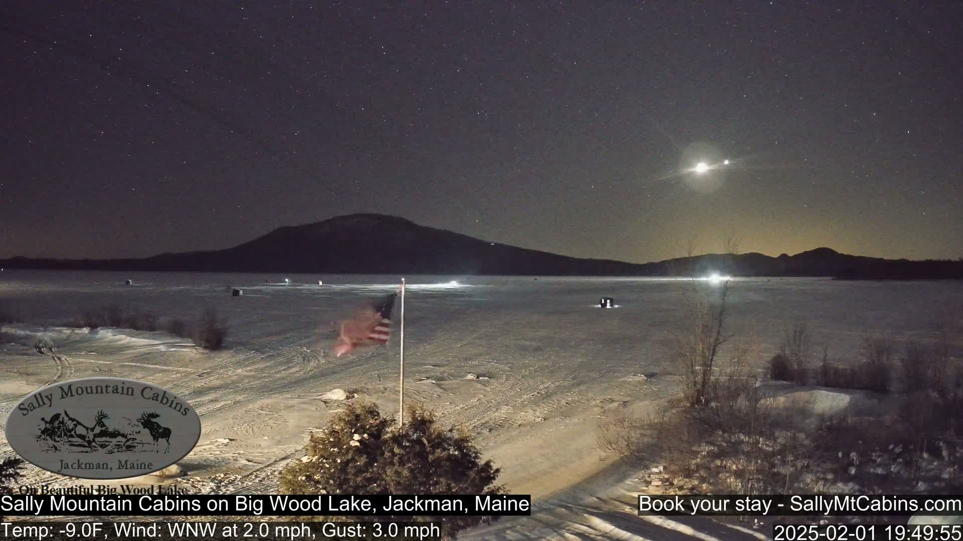 A frozen lake under a starry night sky, with a few lights visible on the ice and a distant mountain range, features an American flag in the foreground.