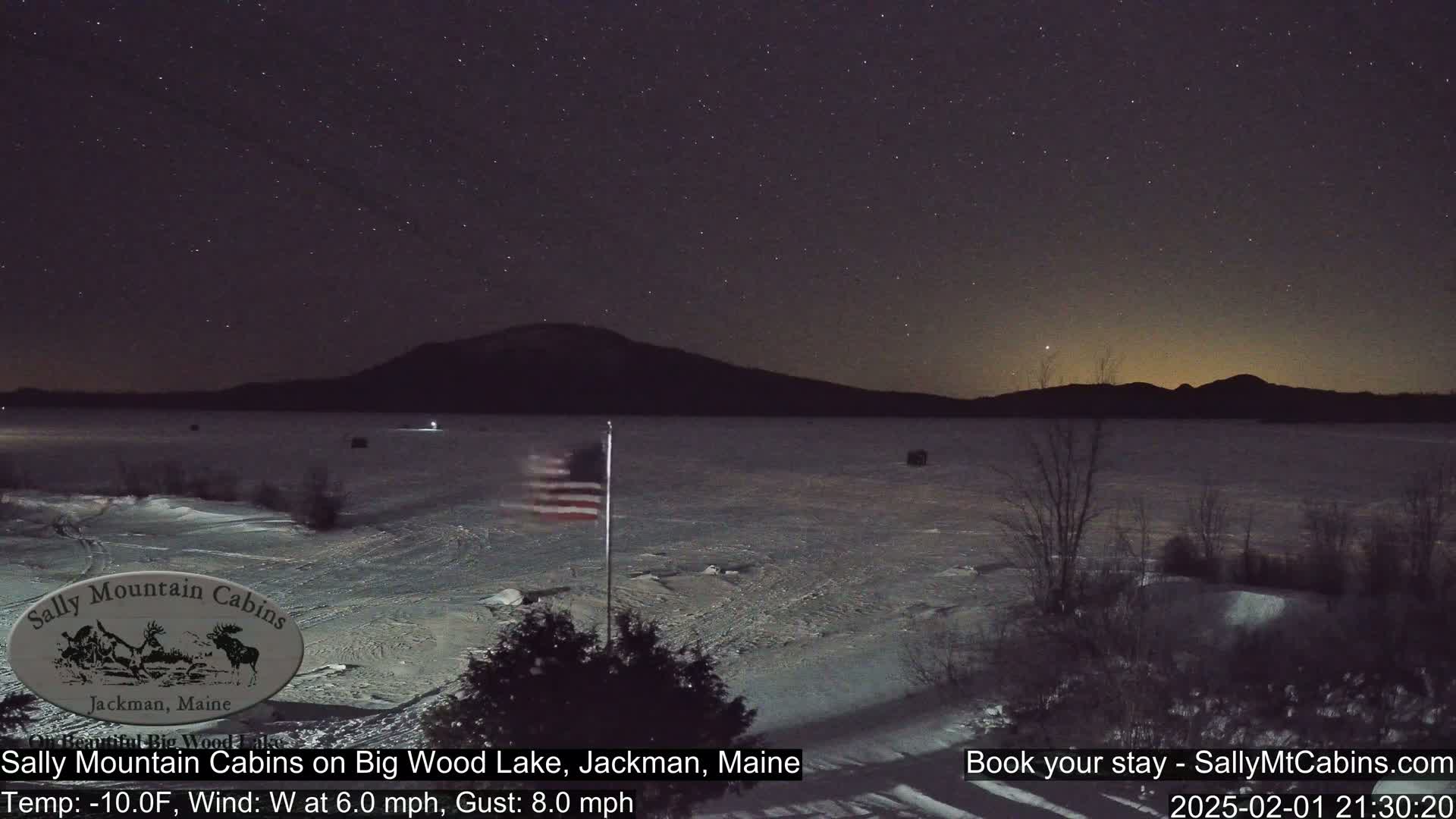A snow-covered lake under a starry night sky, with a distant mountain range and an American flag waving in a 6 mph westerly wind with 8 mph gusts at -10°F.
