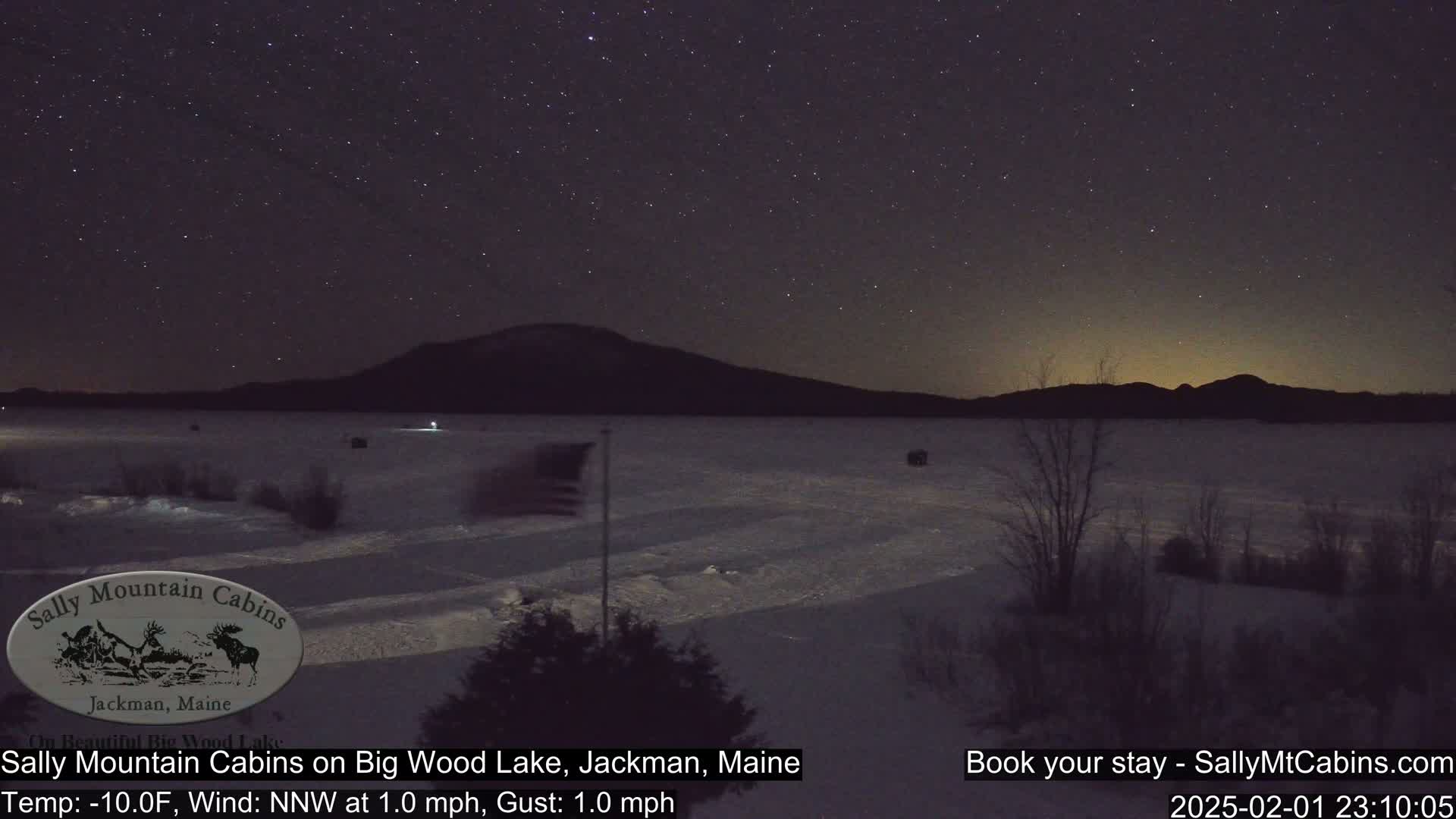 A snow-covered lake is seen at night under a starry sky, with a dark mountain range in the background and a flagpole with an American flag in the foreground.