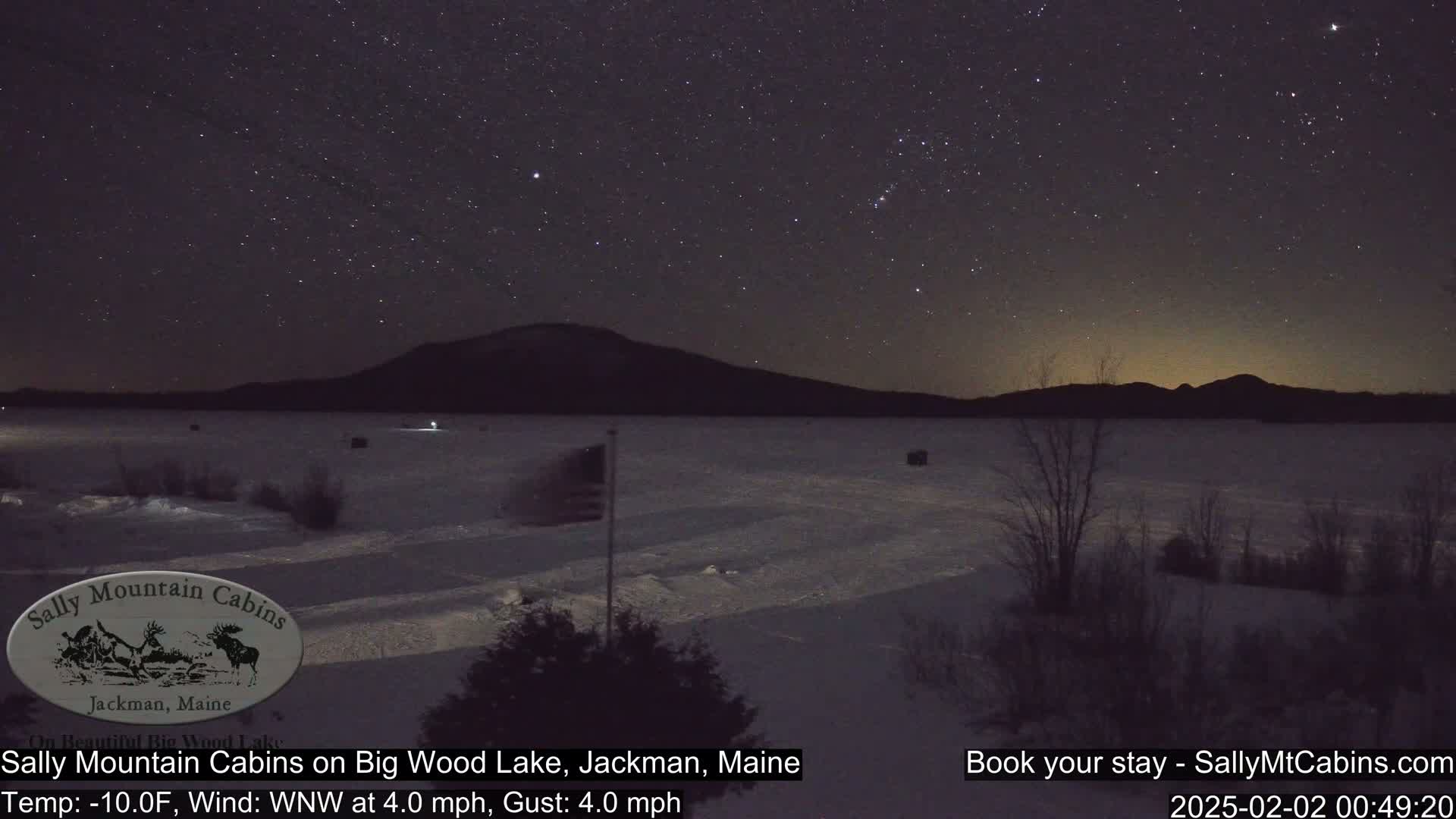 A starry night sky over a snow-covered frozen lake with a dark mountain range in the background.
