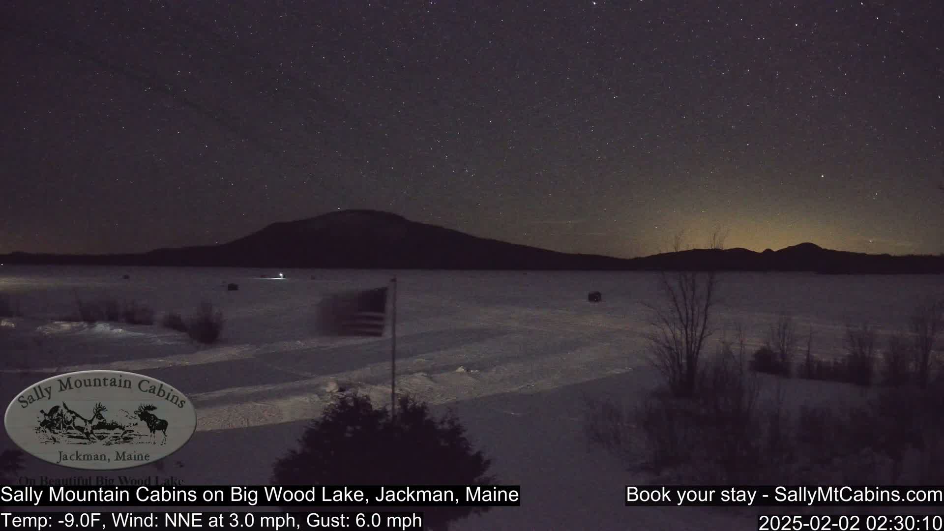 A snow-covered lake is visible under a night sky filled with stars, with a dark mountain range in the background and a flagpole with an American flag in the foreground.