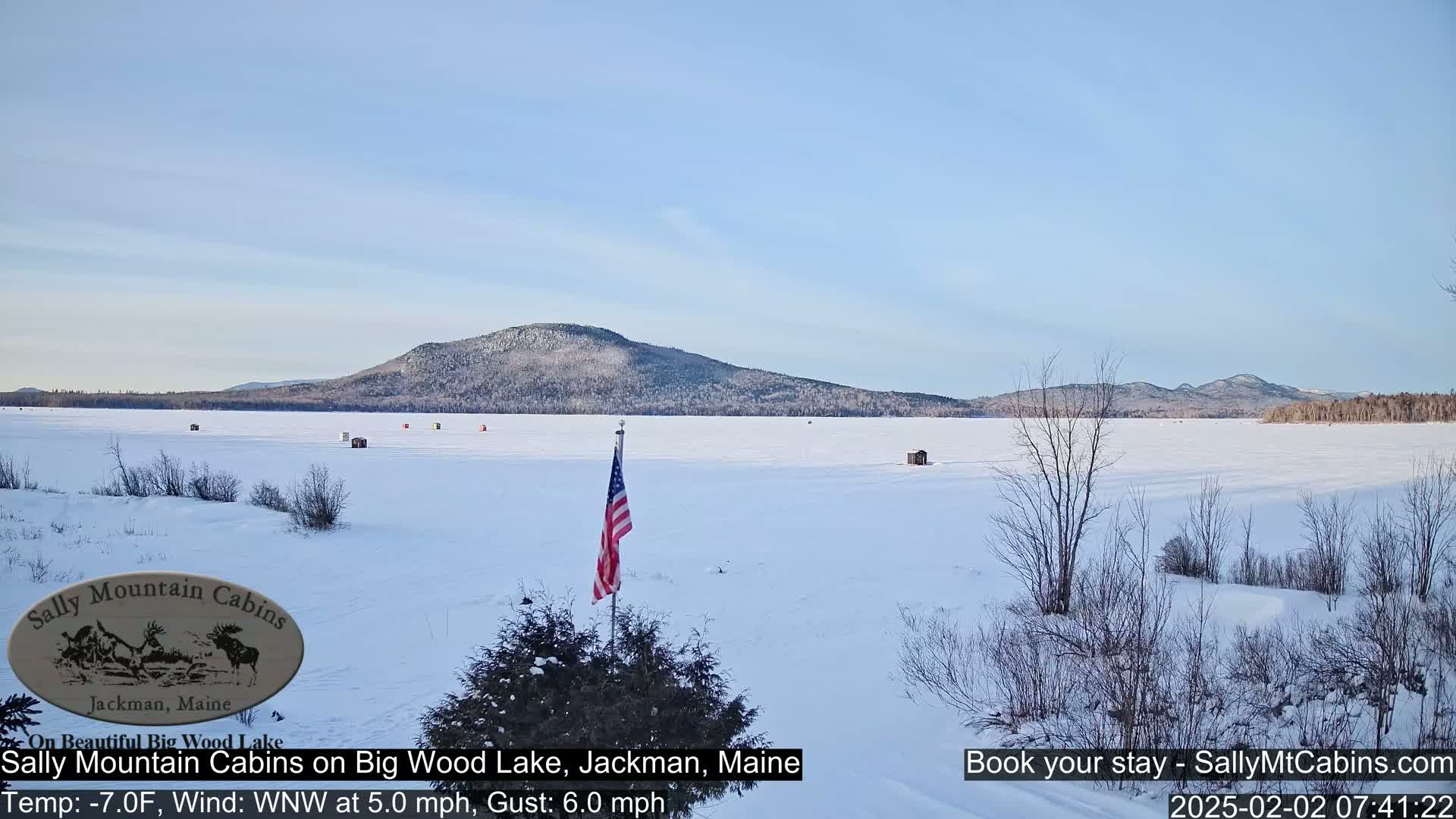 A frozen lake stretches out to a snow-covered mountain range under a clear, light blue sky, with several small ice fishing shelters scattered across the ice and an American flag planted near the foreground.
