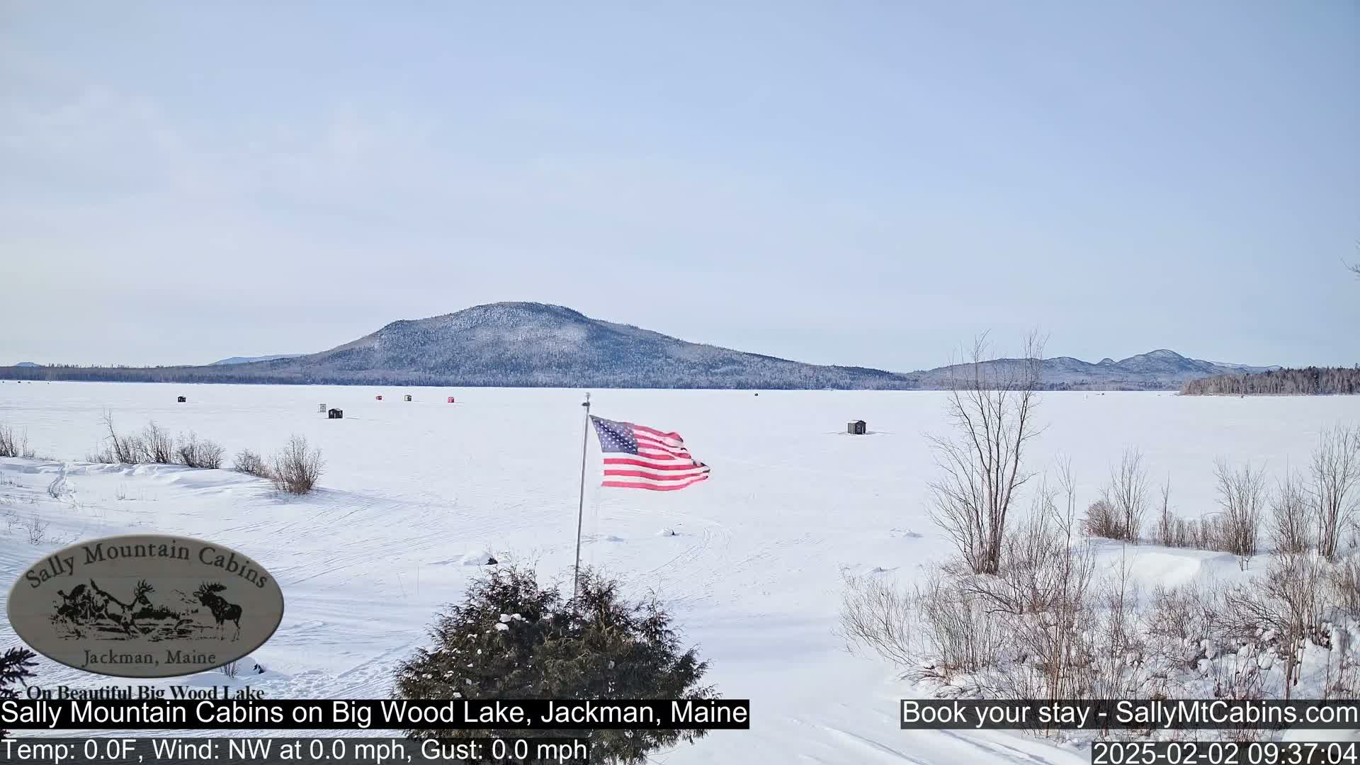 A frozen lake with several ice fishing shacks is seen under a clear sky, with a distant mountain range and an American flag waving in the foreground.