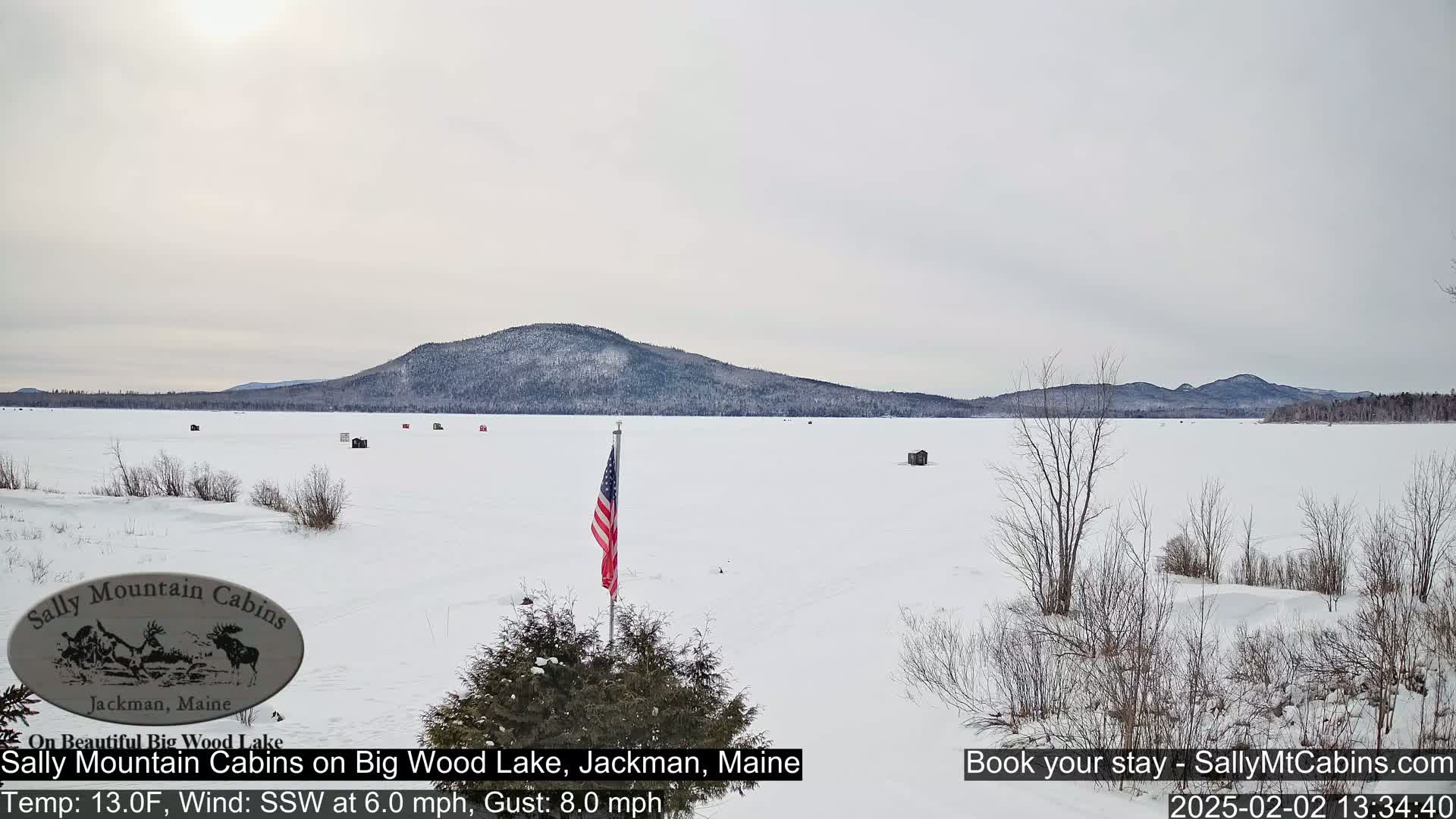 A frozen lake stretches to snow-covered mountains under a cloudy sky, with a few ice fishing shacks and an American flag visible.