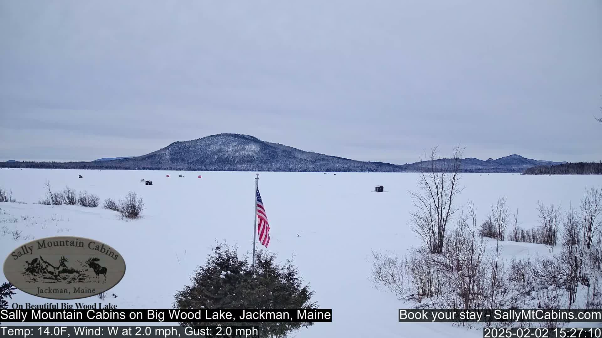 A frozen lake with several small ice fishing huts, a distant snow-covered mountain range, and an American flag is visible under an overcast sky.
