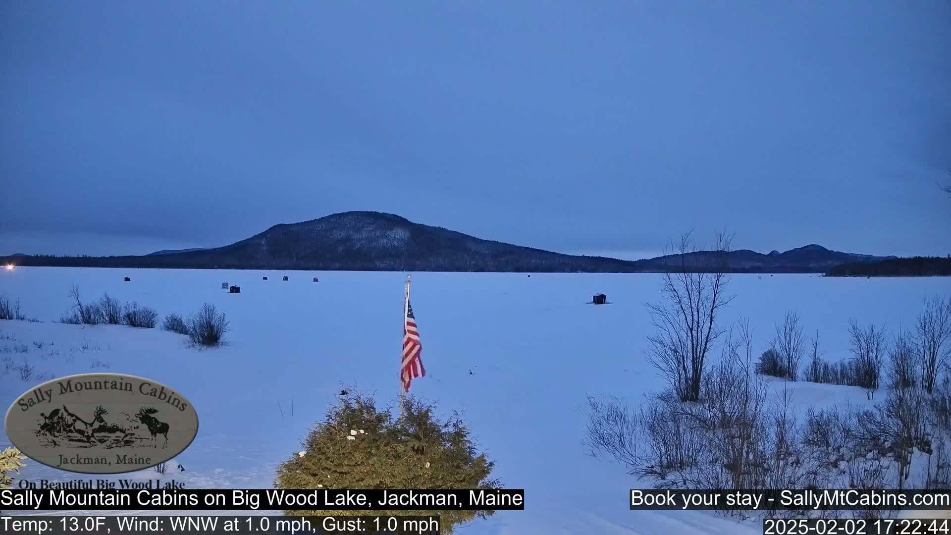 A frozen lake under a twilight sky is seen from a snowy shoreline, with a few ice fishing huts scattered across the ice and a US flag planted in the foreground.