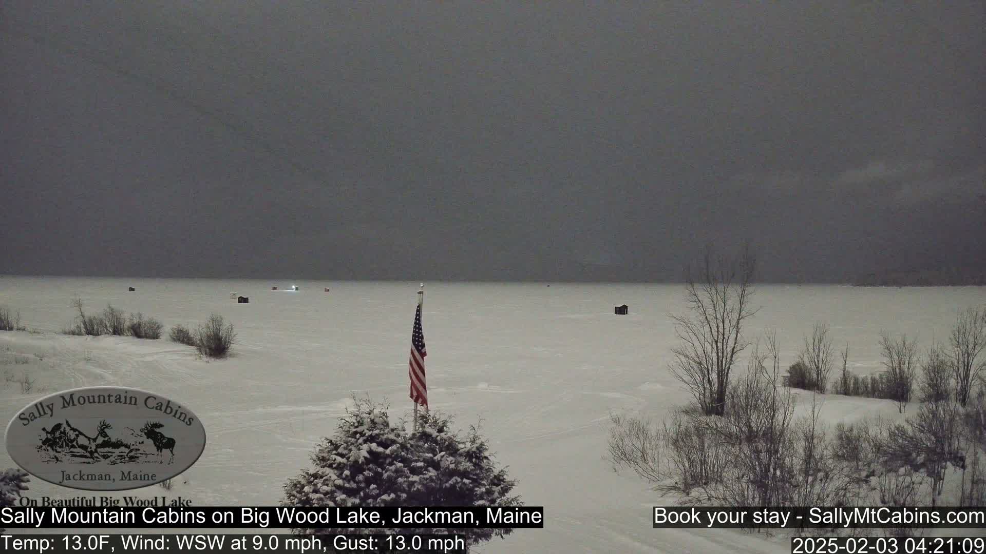 A snow-covered, frozen lake under a dark, overcast sky, with several small structures and an American flag visible in the foreground.