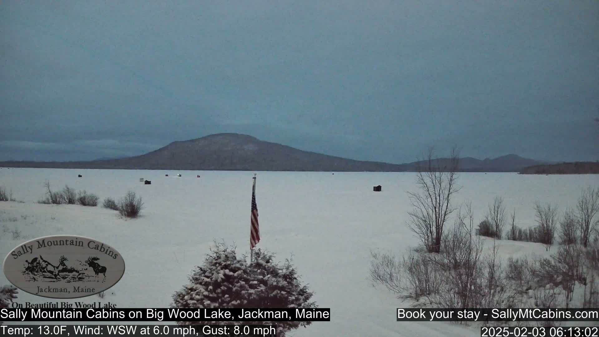 A snow-covered frozen lake stretches to a distant mountain range under a gray sky, with a few ice fishing shelters scattered across the ice and a small American flag in the foreground.