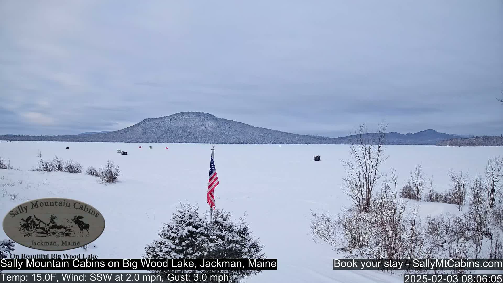 A snow-covered frozen lake with a few ice fishing shacks and an American flag is seen, with a snow-covered mountain range in the background under a cloudy sky.