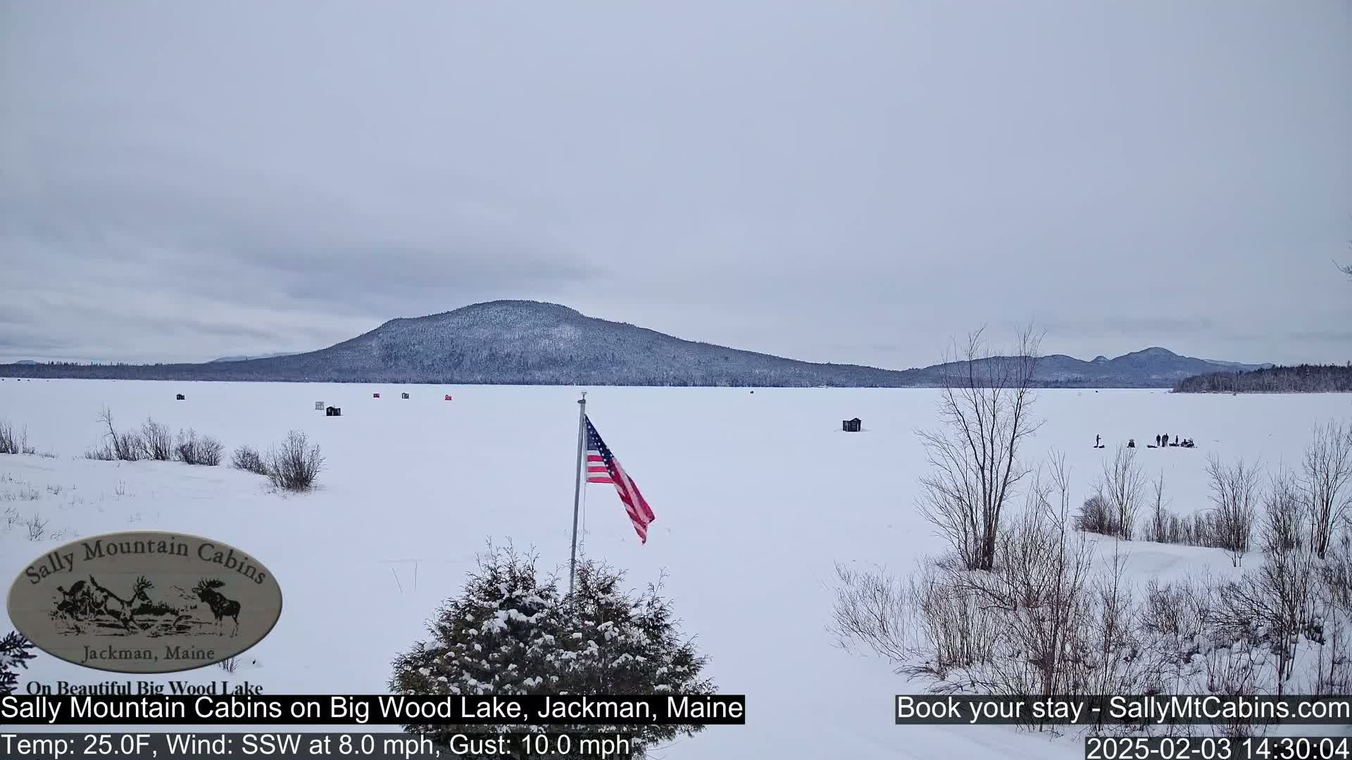 A snow-covered frozen lake stretches to a distant mountain range under an overcast sky, with a few ice fishing shelters and a US flag visible.