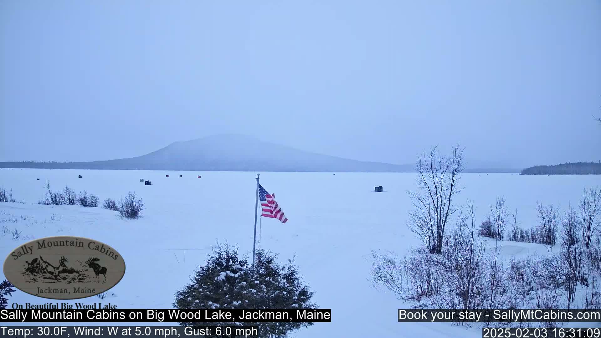 A snow-covered frozen lake stretches to a distant mountain range under an overcast sky, with a few ice fishing huts scattered across the ice and an American flag waving in the wind.