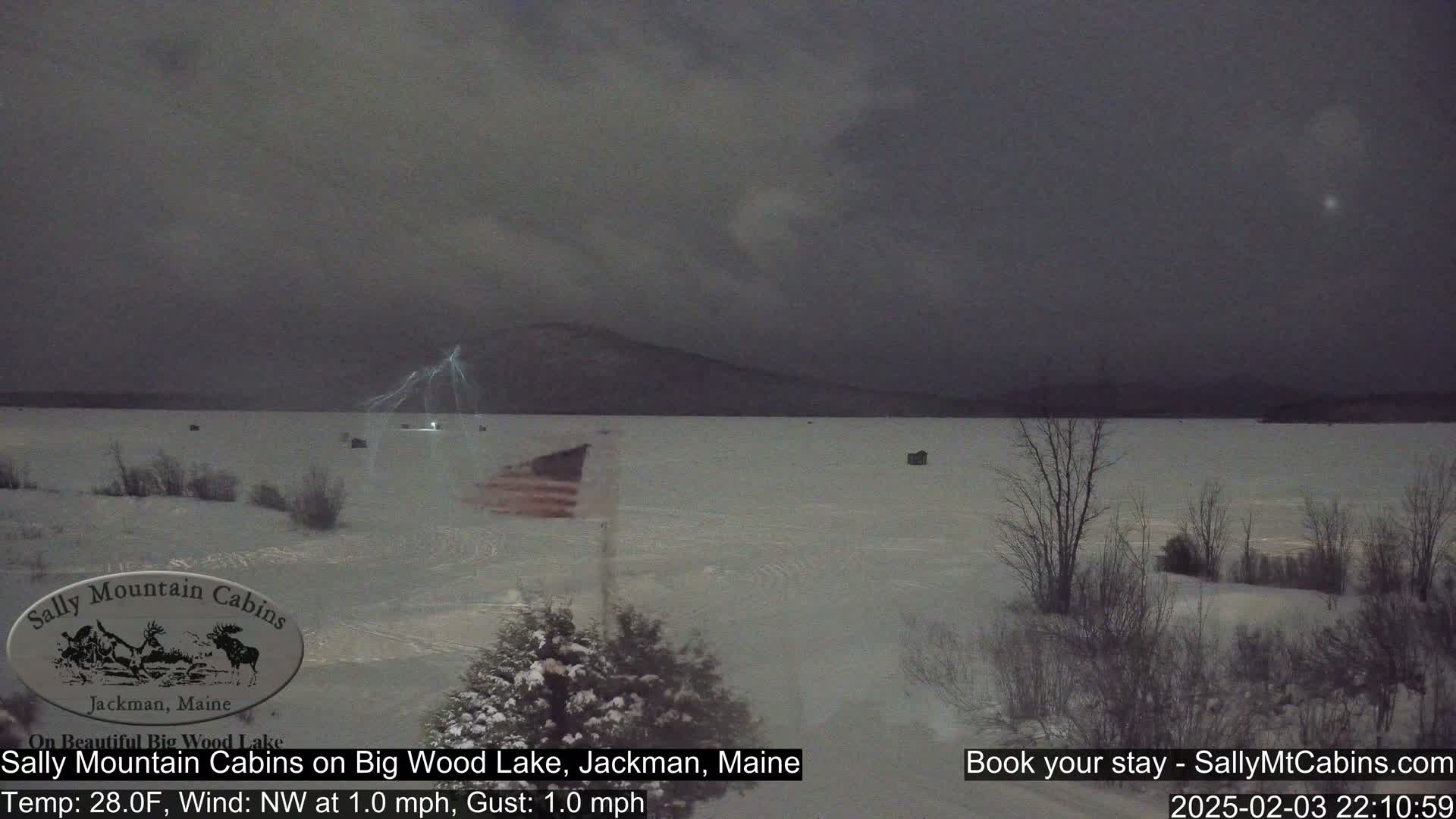 A snow-covered, frozen lake is shown at night under a dark, stormy sky with a lightning strike visible in the distance; a small American flag is planted in the foreground.