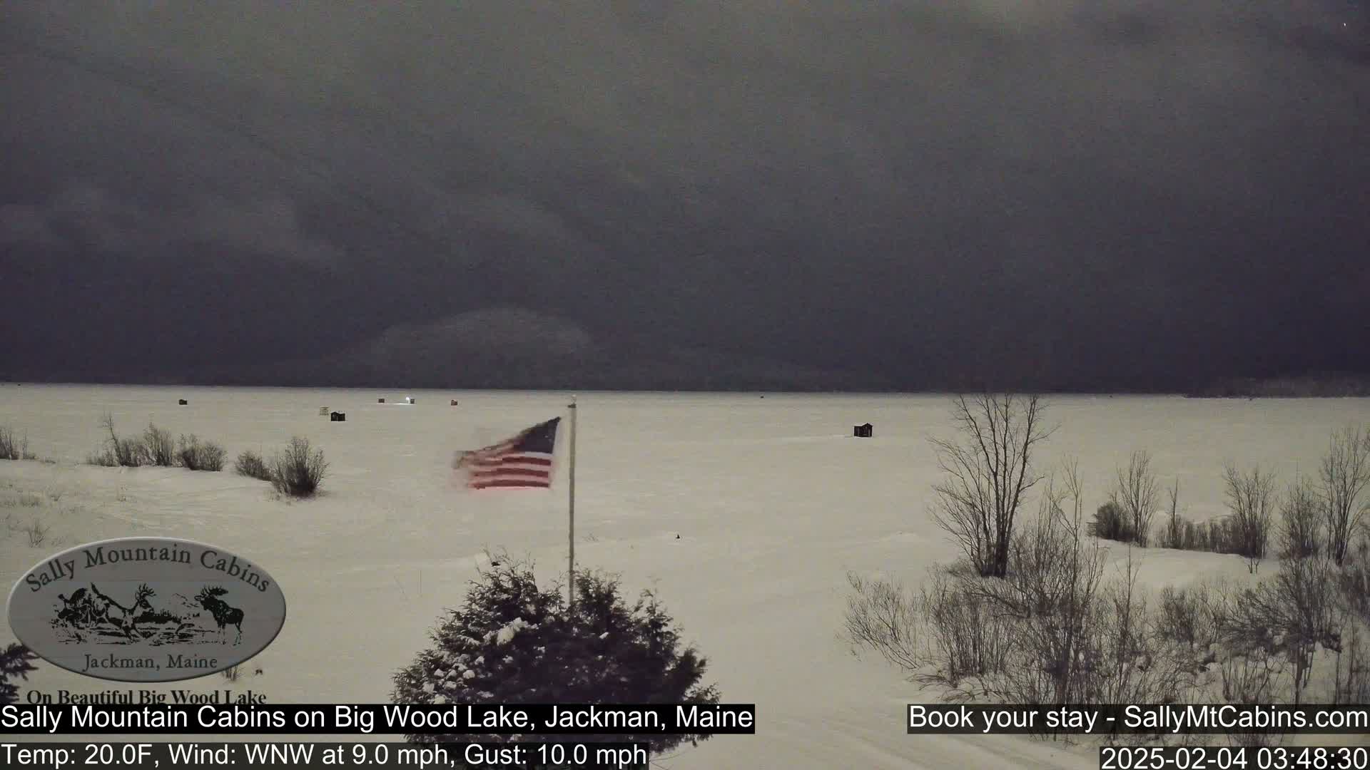 A snow-covered frozen lake under a dark, overcast sky with an American flag waving in the foreground.