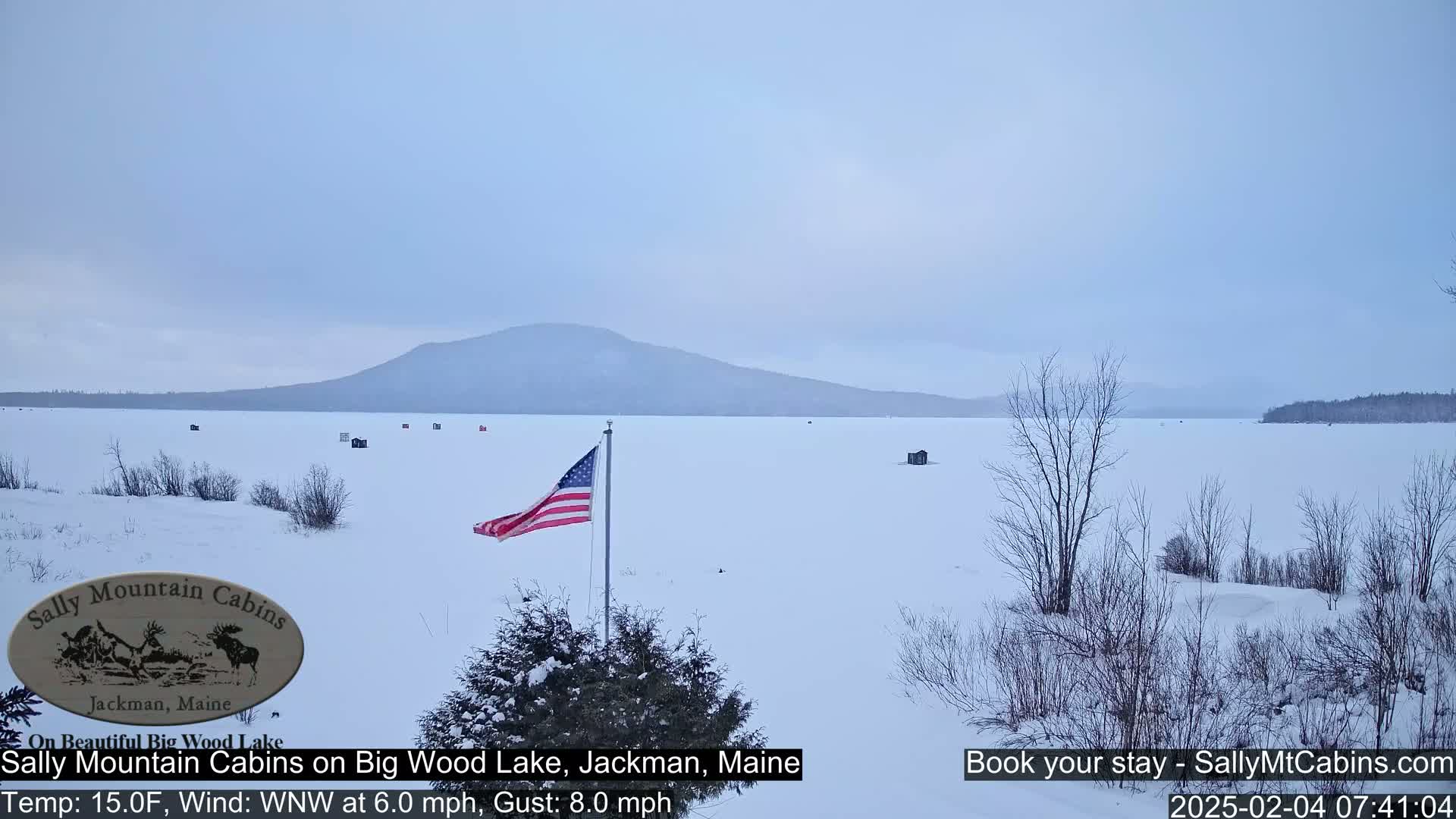 A snow-covered frozen lake stretches out to a distant mountain range under a pale, overcast sky, with a few ice fishing shacks and an American flag visible in the foreground.