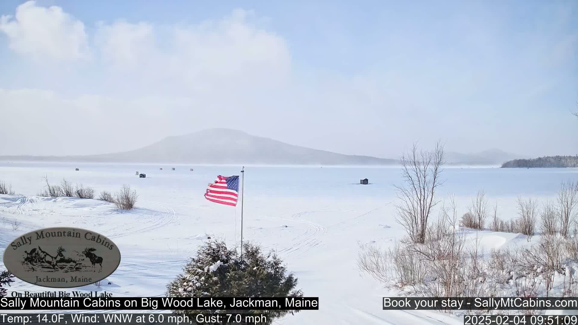 A snow-covered frozen lake stretches to a hazy mountain range under a partly cloudy sky, with an American flag waving near the foreground.