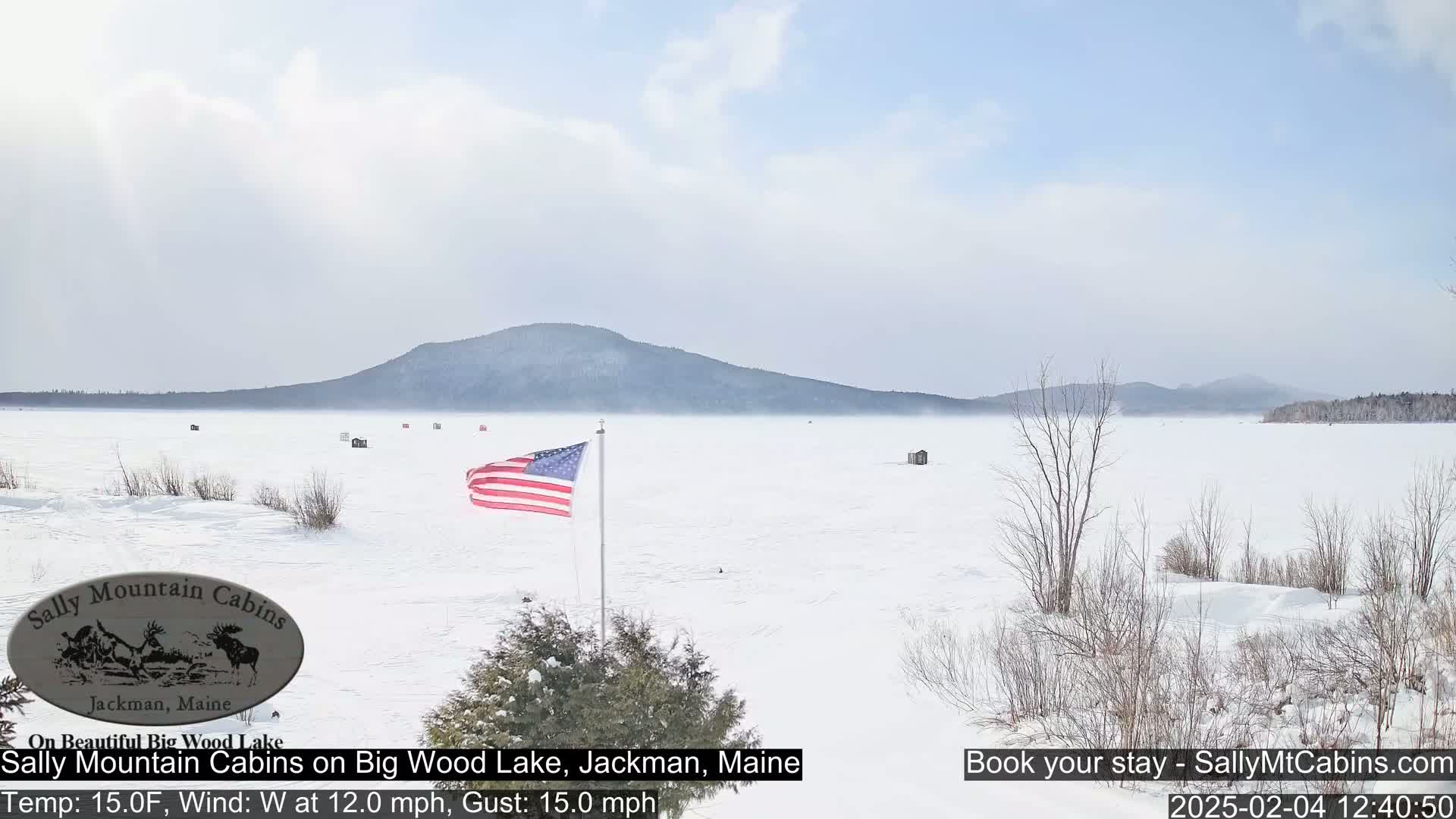 A snow-covered frozen lake stretches to a distant mountain range under a mostly clear sky, with a few ice fishing shelters scattered across the ice and an American flag waving in the west wind.