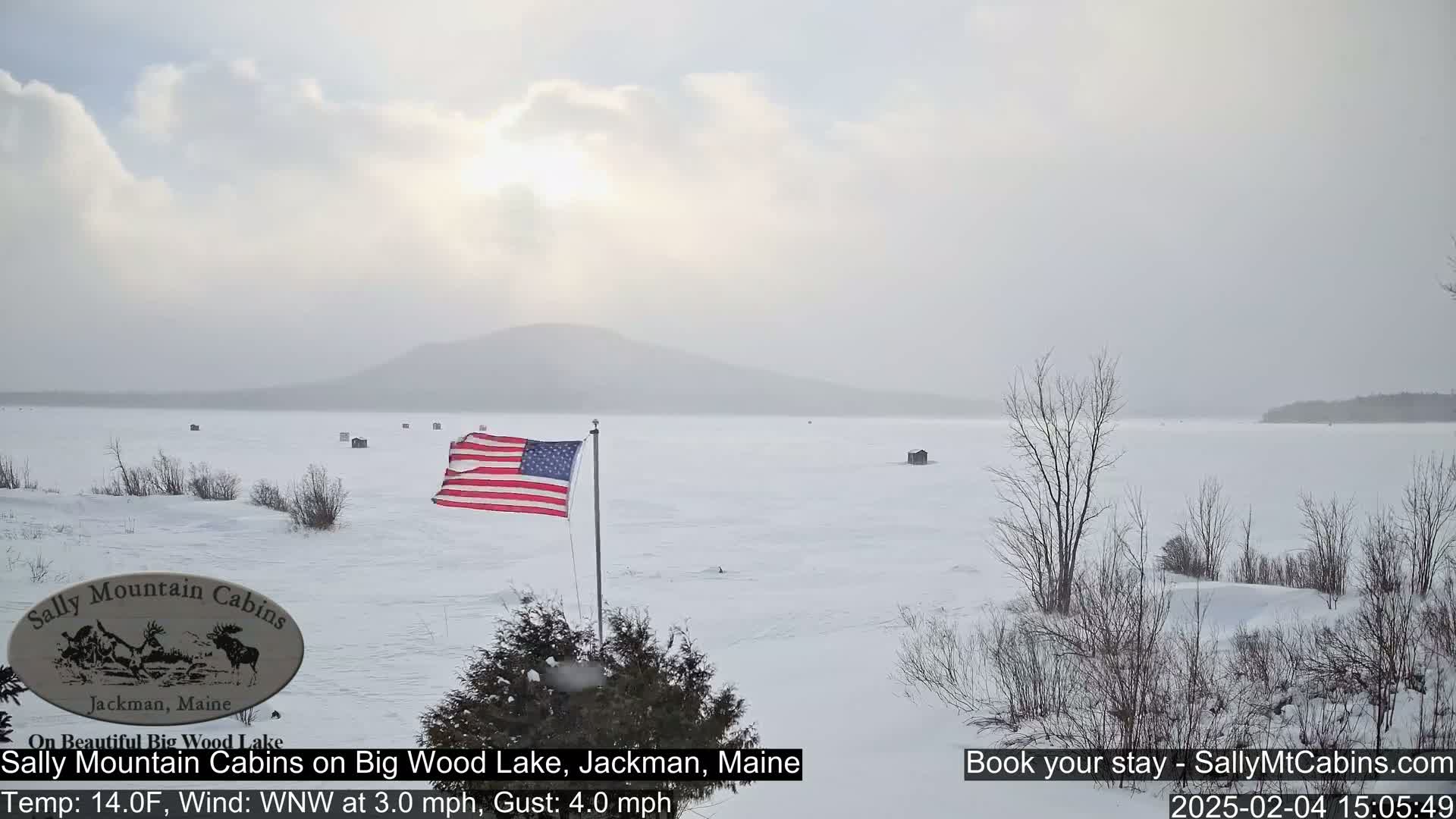 A frozen lake under a hazy sky, with a distant mountain range and an American flag visible near the foreground, amidst a snowy landscape.