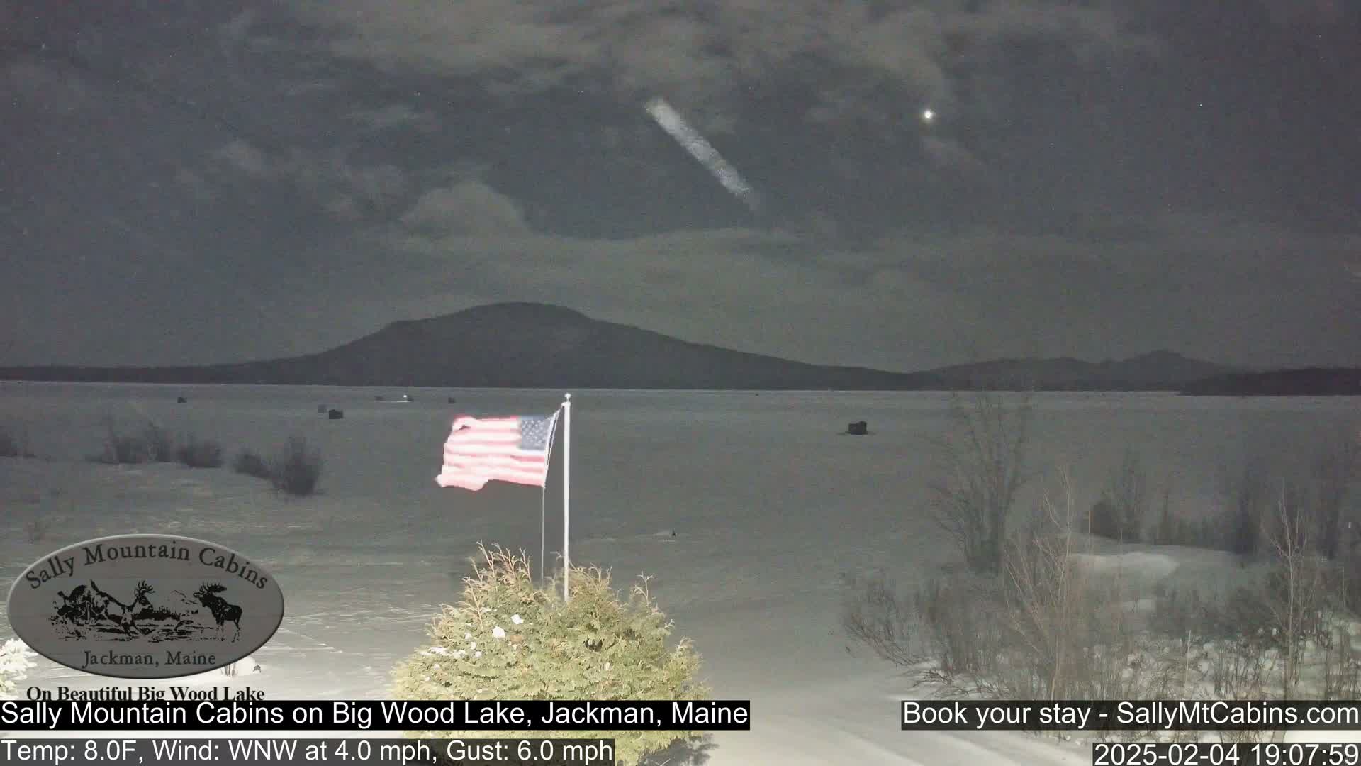 A nighttime view of a frozen lake under a cloudy sky shows a distant mountain range, an American flag on a pole, and some sparse vegetation in the foreground.