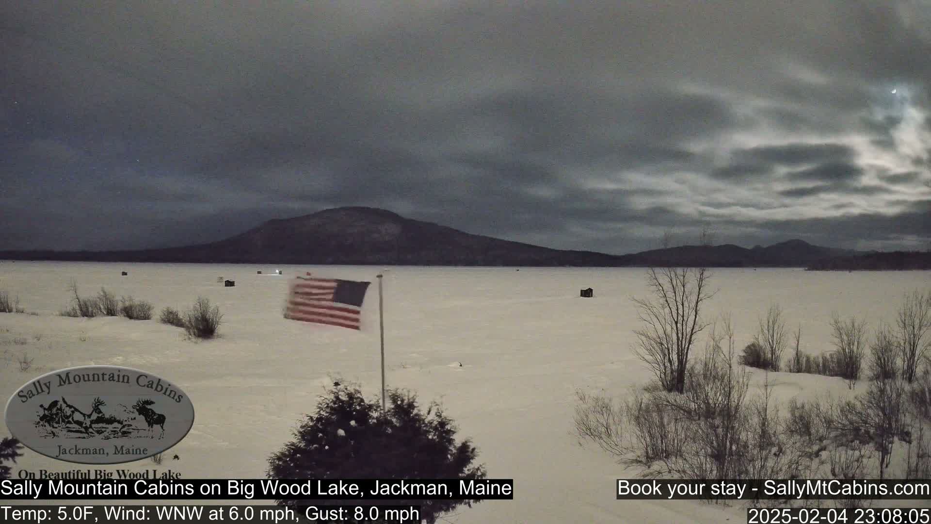 A snow-covered frozen lake stretches out to a dark mountain range under a cloudy night sky.