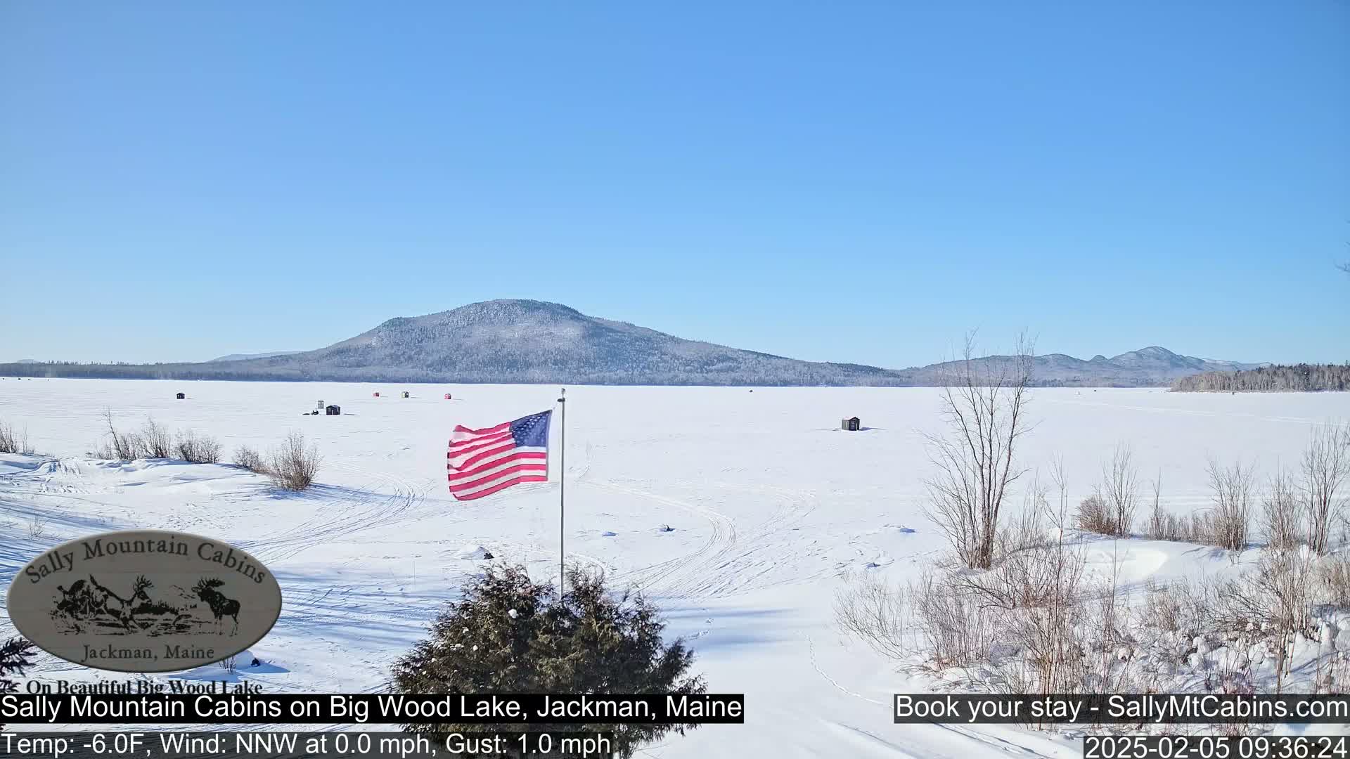 A frozen lake, under a clear blue sky, stretches towards a distant mountain range, with a few ice fishing shacks scattered across its surface and an American flag waving near the foreground.