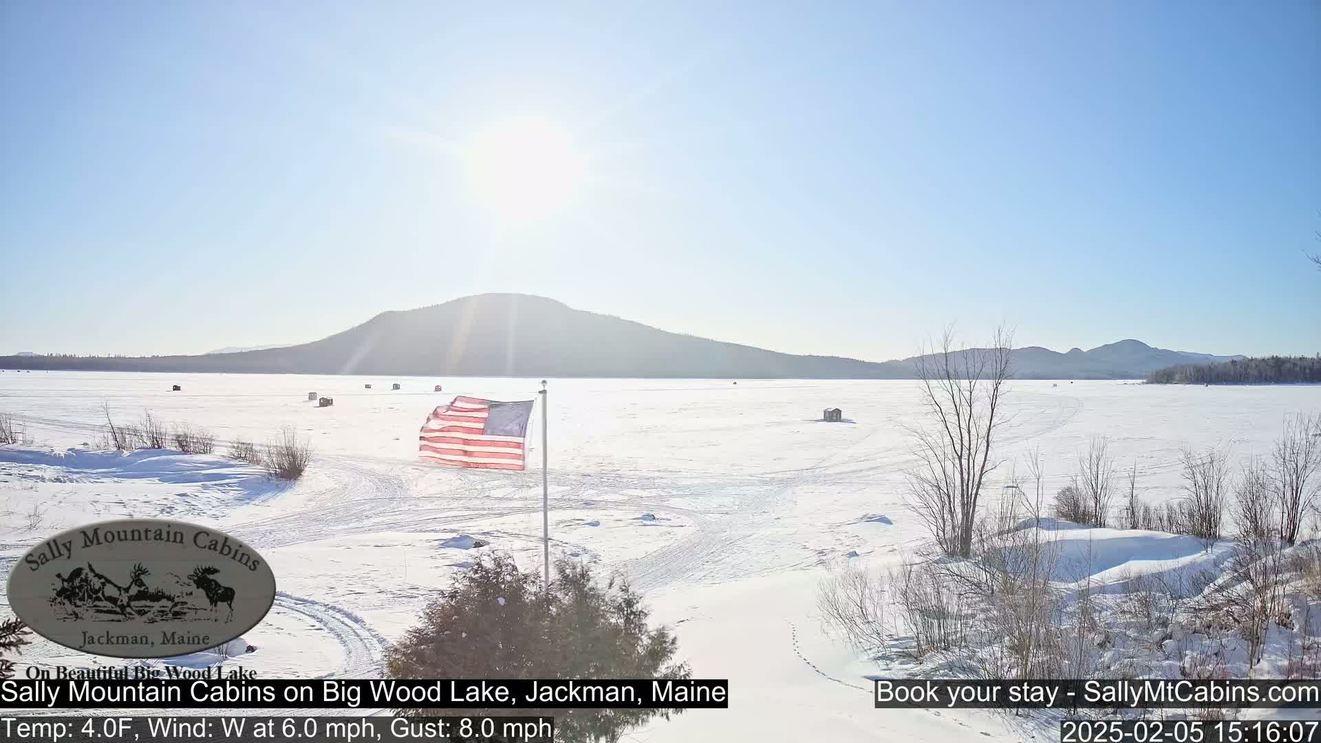 A frozen lake stretches to a distant mountain range under a clear blue sky, with an American flag waving near the foreground and several small structures scattered across the ice.