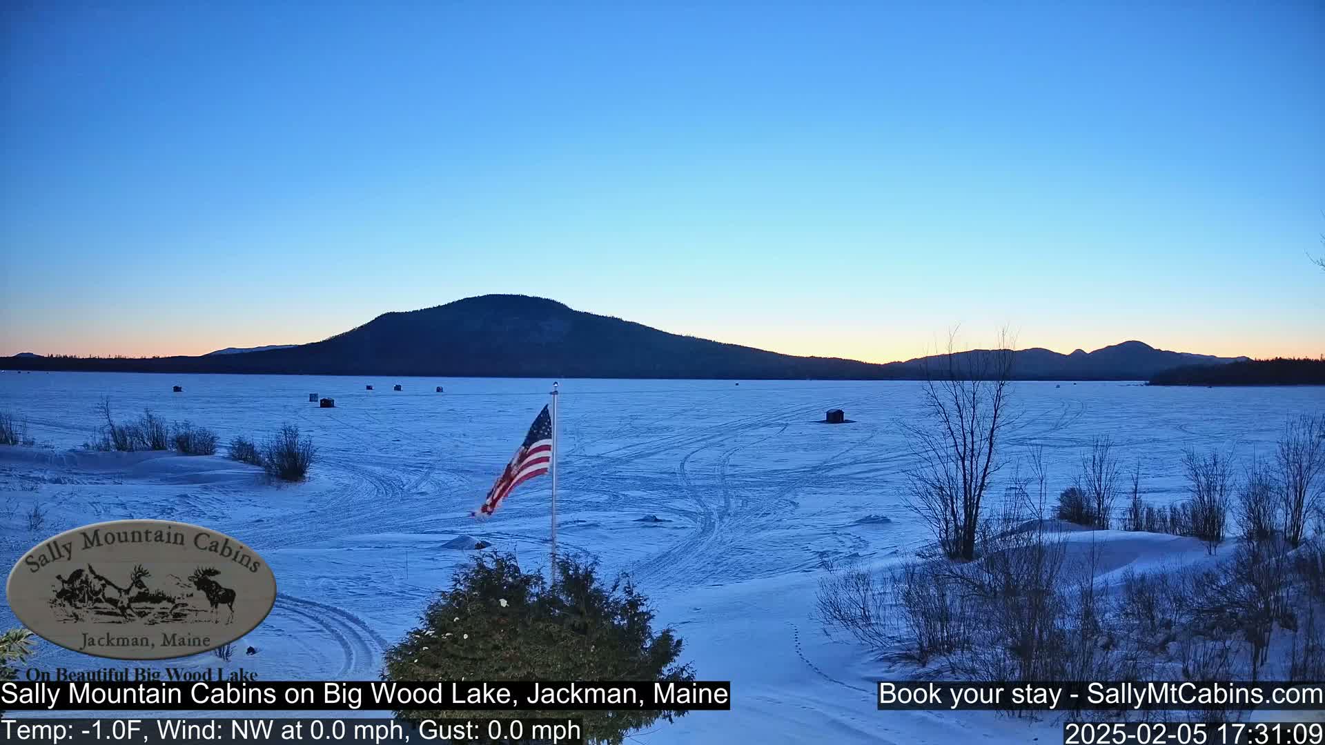A frozen lake stretches out before a snow-covered shoreline, featuring a few ice fishing huts and an American flag, all under a twilight sky.