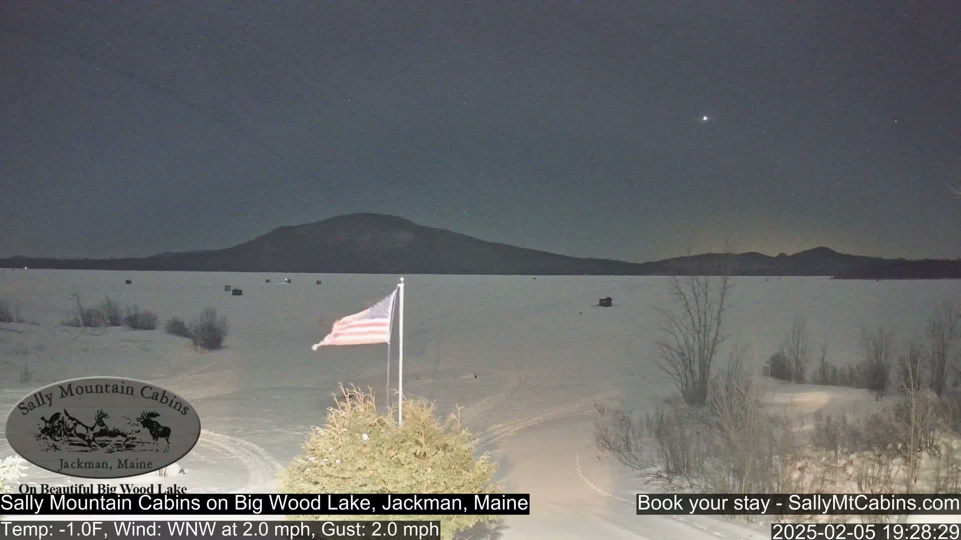 A frozen lake at night is viewed from a shoreline under a clear sky with a single bright star visible, and an American flag waves gently in the wind near a stand of evergreens.