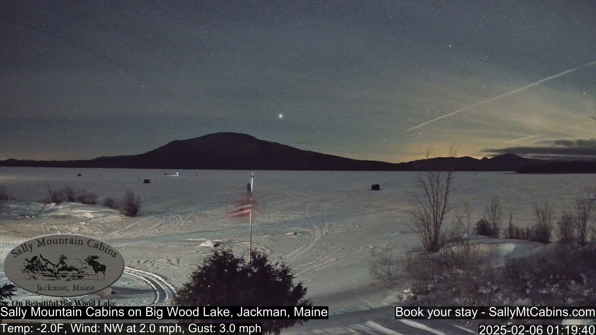 A frozen lake under a starry night sky, with a mountain in the background and a US flag on the shore, in cold, windy conditions.