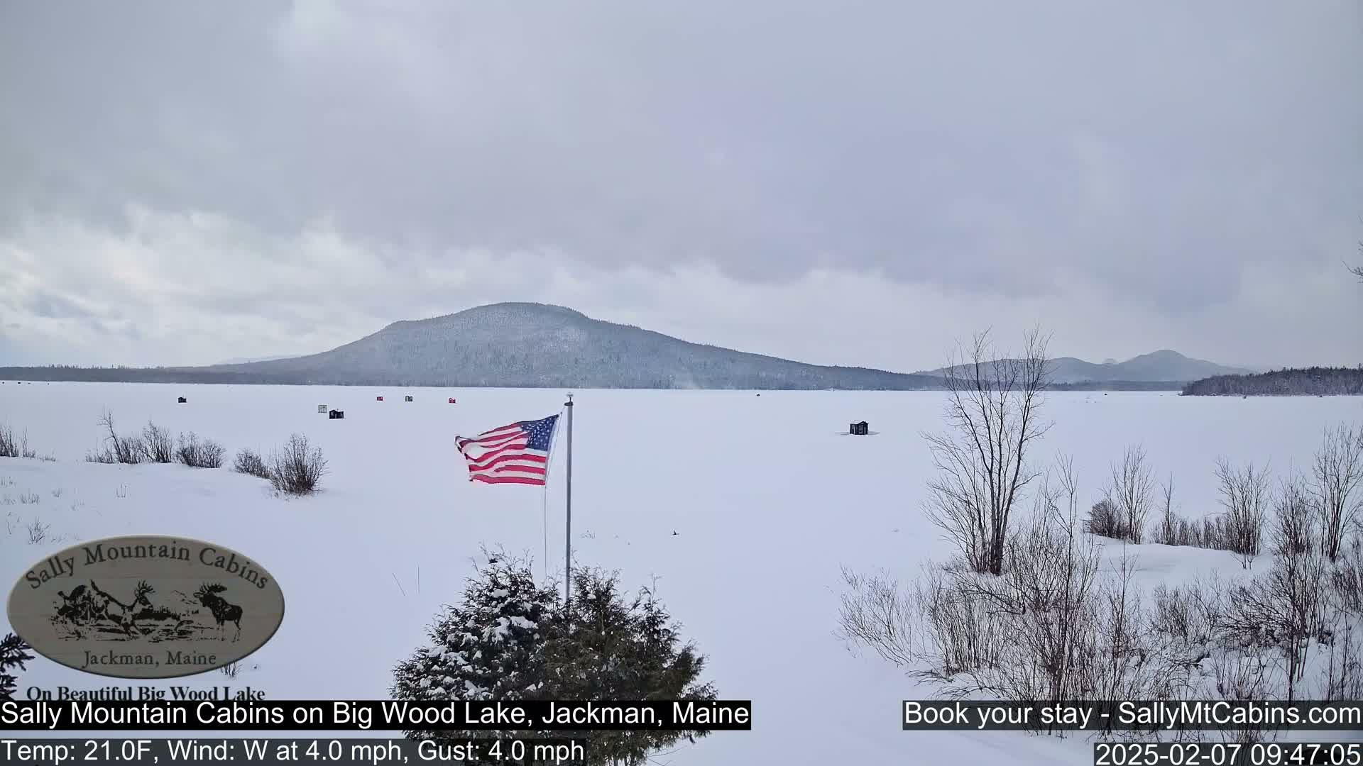 A snow-covered frozen lake stretches to a snow-dusted mountain range under an overcast sky, with a few ice fishing huts and an American flag visible.