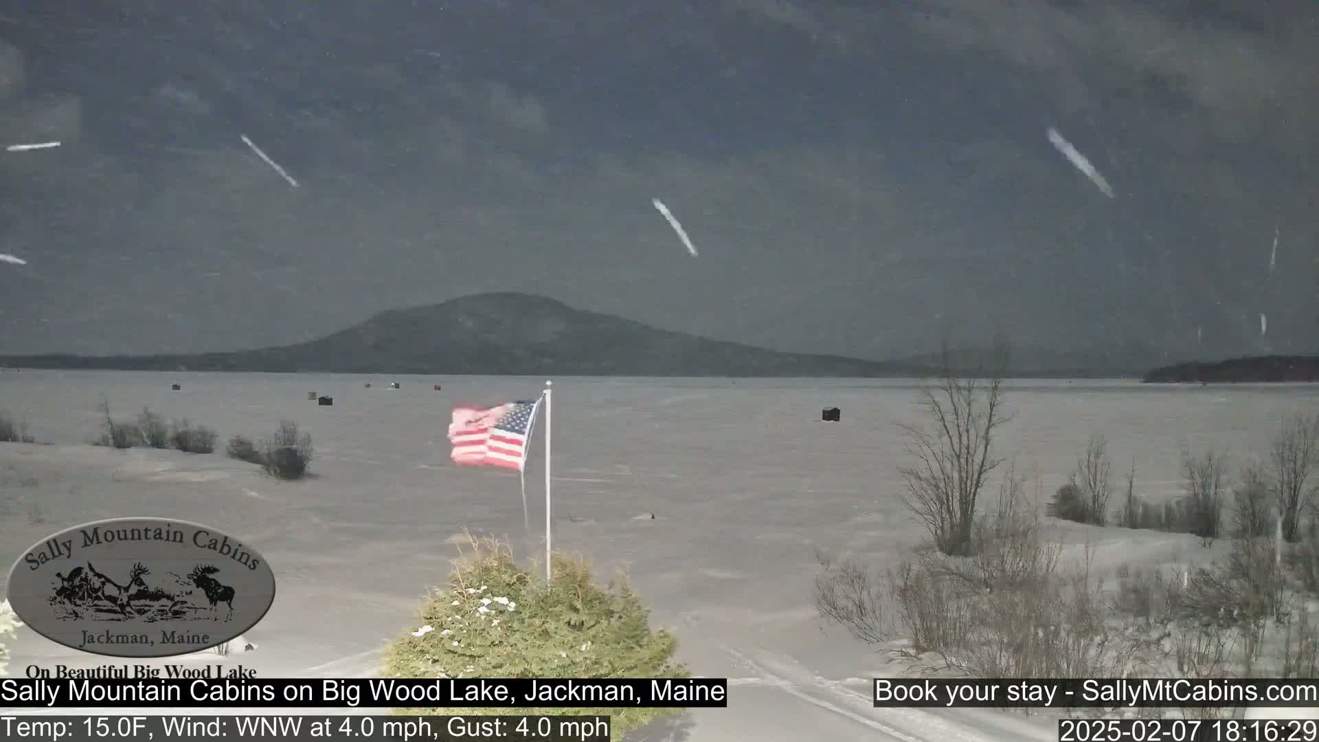 A snow-covered frozen lake stretches to a distant mountain range under a lightly snowing, dark sky, with an American flag waving in the foreground.