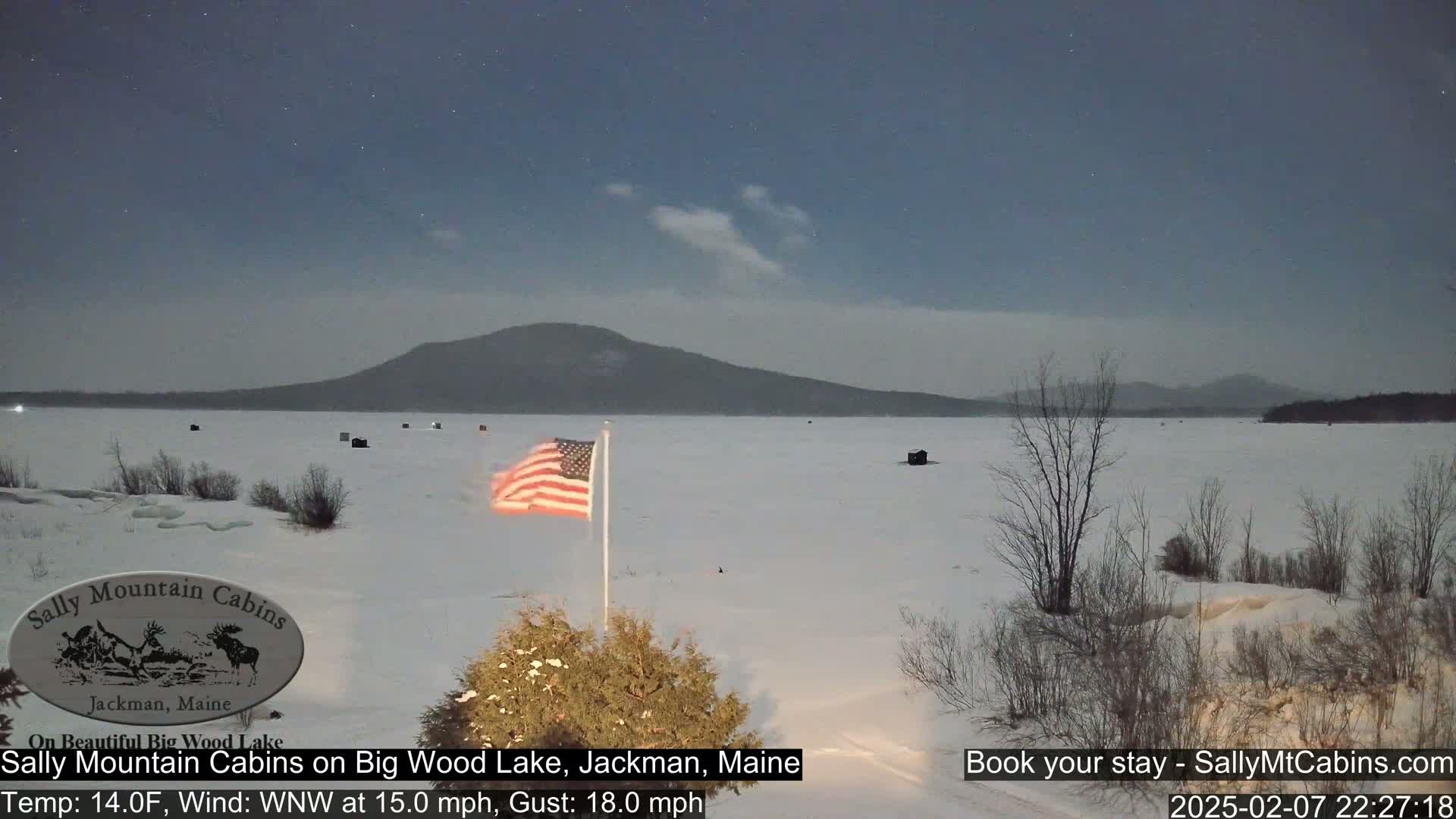 A frozen lake under a mostly clear night sky, with a few clouds and stars visible, features a snow-covered shoreline, some ice fishing huts, and an American flag waving in a WNW wind.