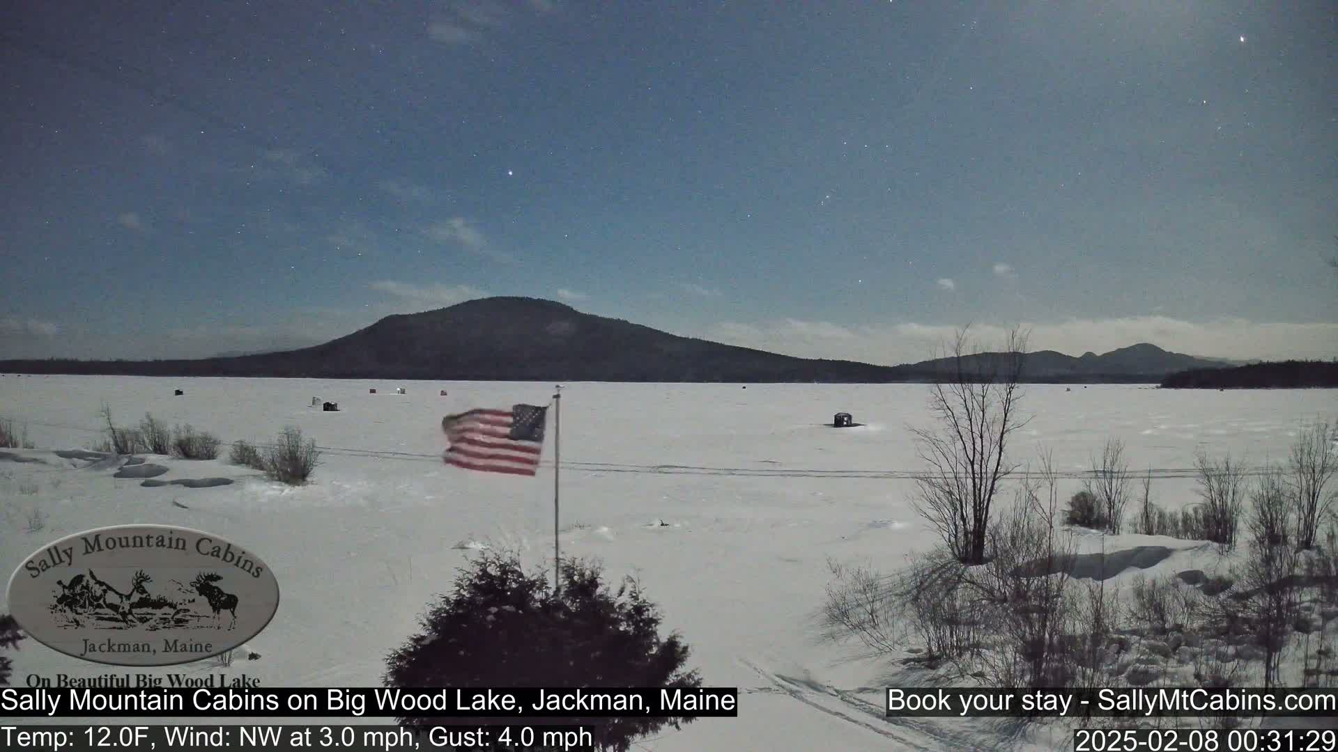 A snow-covered frozen lake is seen under a clear night sky with stars visible, a US flag waves gently in the breeze near a shoreline with sparse, snow-covered vegetation, and distant mountains are visible in the background.
