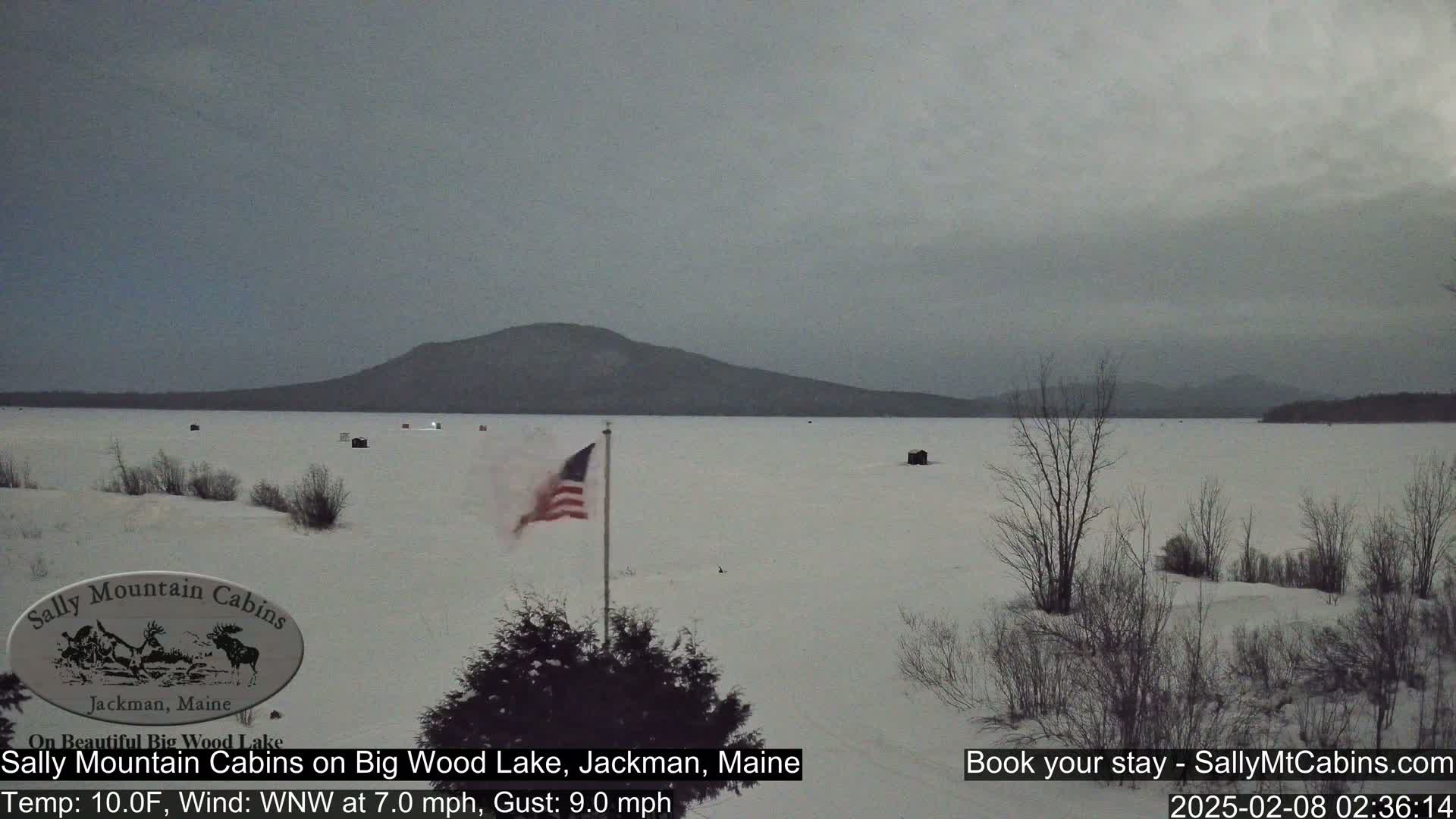 A snow-covered frozen lake stretches towards a distant mountain range under a cloudy sky, with a few ice fishing huts visible and an American flag waving in the wind.