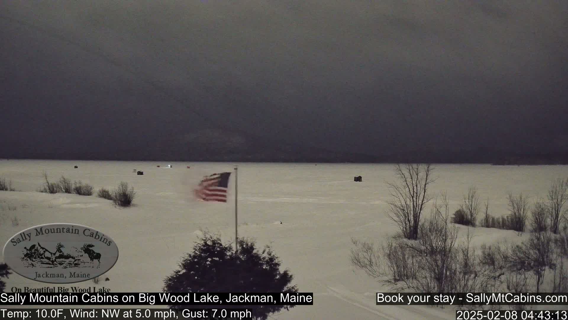 A snow-covered frozen lake is visible under a dark, overcast sky, with a few small structures and a waving American flag in the foreground.