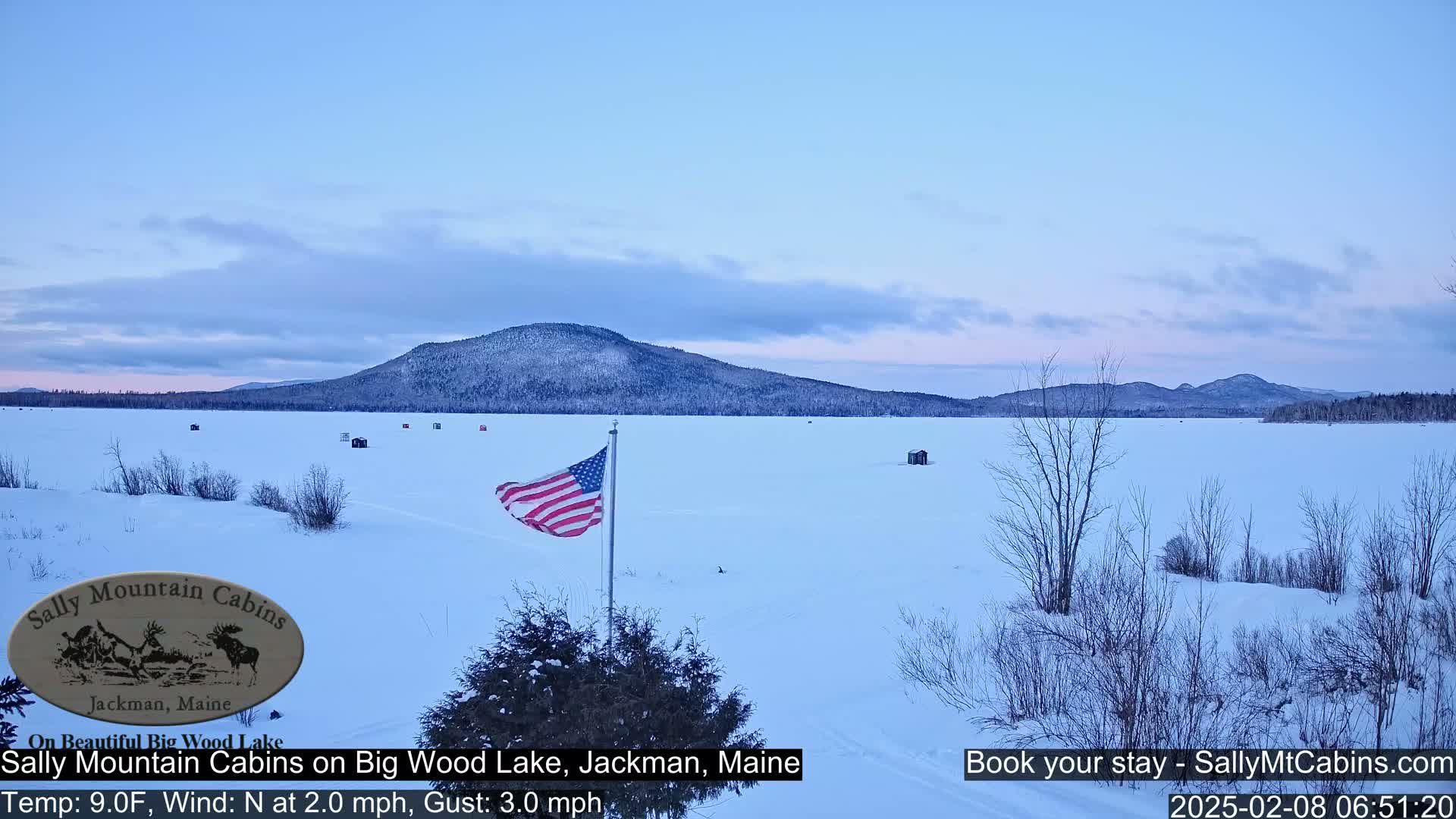 A frozen lake with scattered ice fishing huts is seen under a pale blue sky, with a snow-covered shoreline and a distant, snow-capped mountain range.