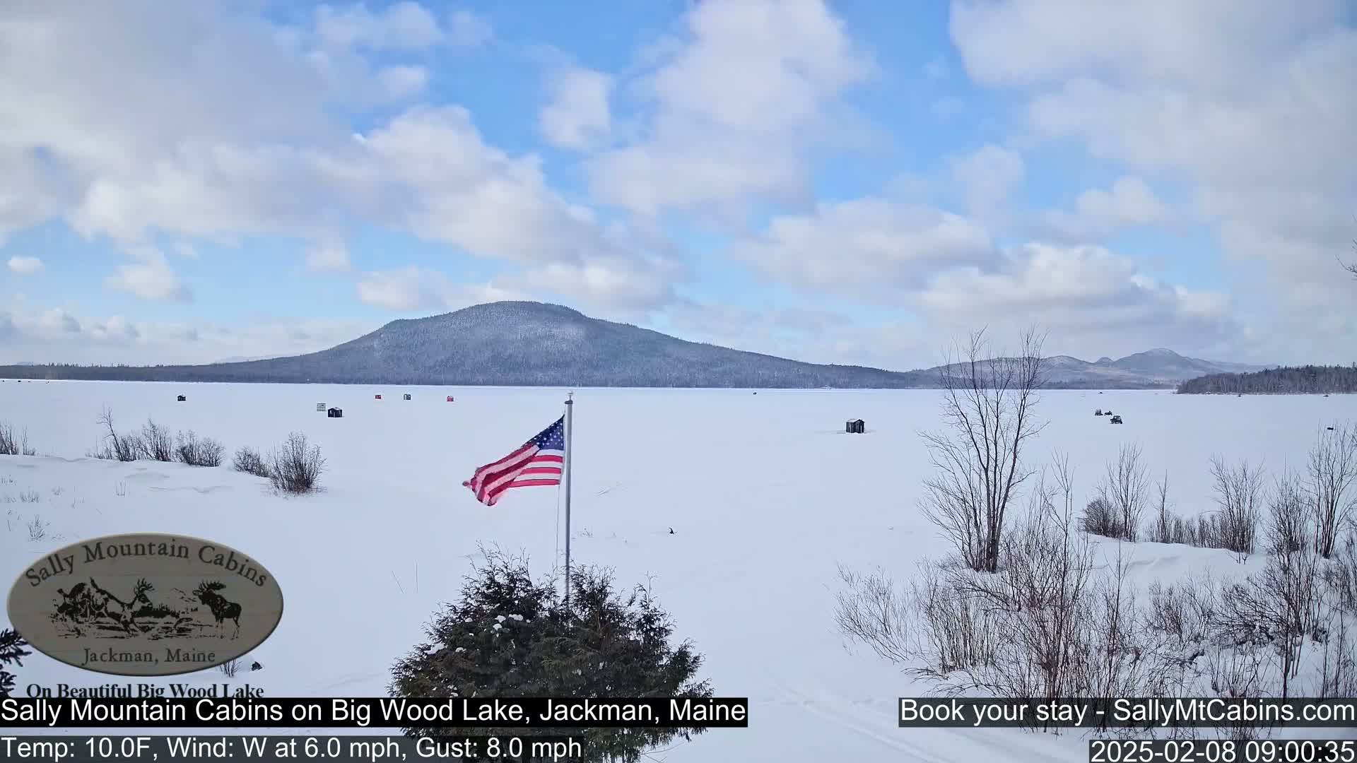 A snow-covered frozen lake stretches to a distant mountain range under a partly cloudy sky, with several ice fishing huts scattered across the ice and an American flag waving in the foreground.