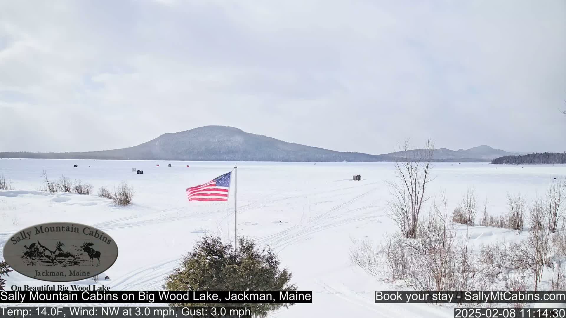 A snow-covered frozen lake stretches to a distant mountain range under a bright, overcast sky, with an American flag and several ice fishing huts visible.