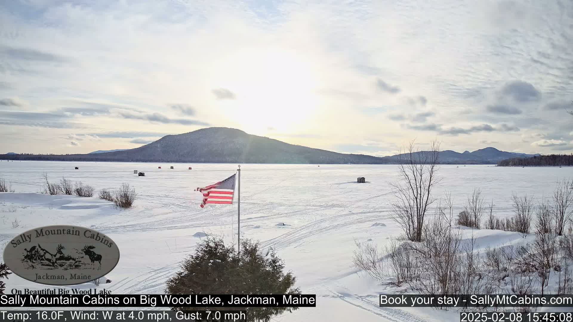 A snow-covered frozen lake stretches towards distant mountains under a partly cloudy sky, with an American flag waving near the foreground.